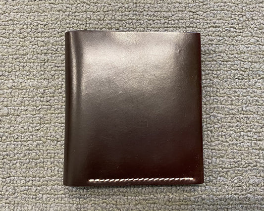 The Alden Madison LG 7008 - Alden Color 8 Shell Cordovan Wallet, closed and dark brown with contrasting white bottom stitching, rests on a textured light gray carpet.
