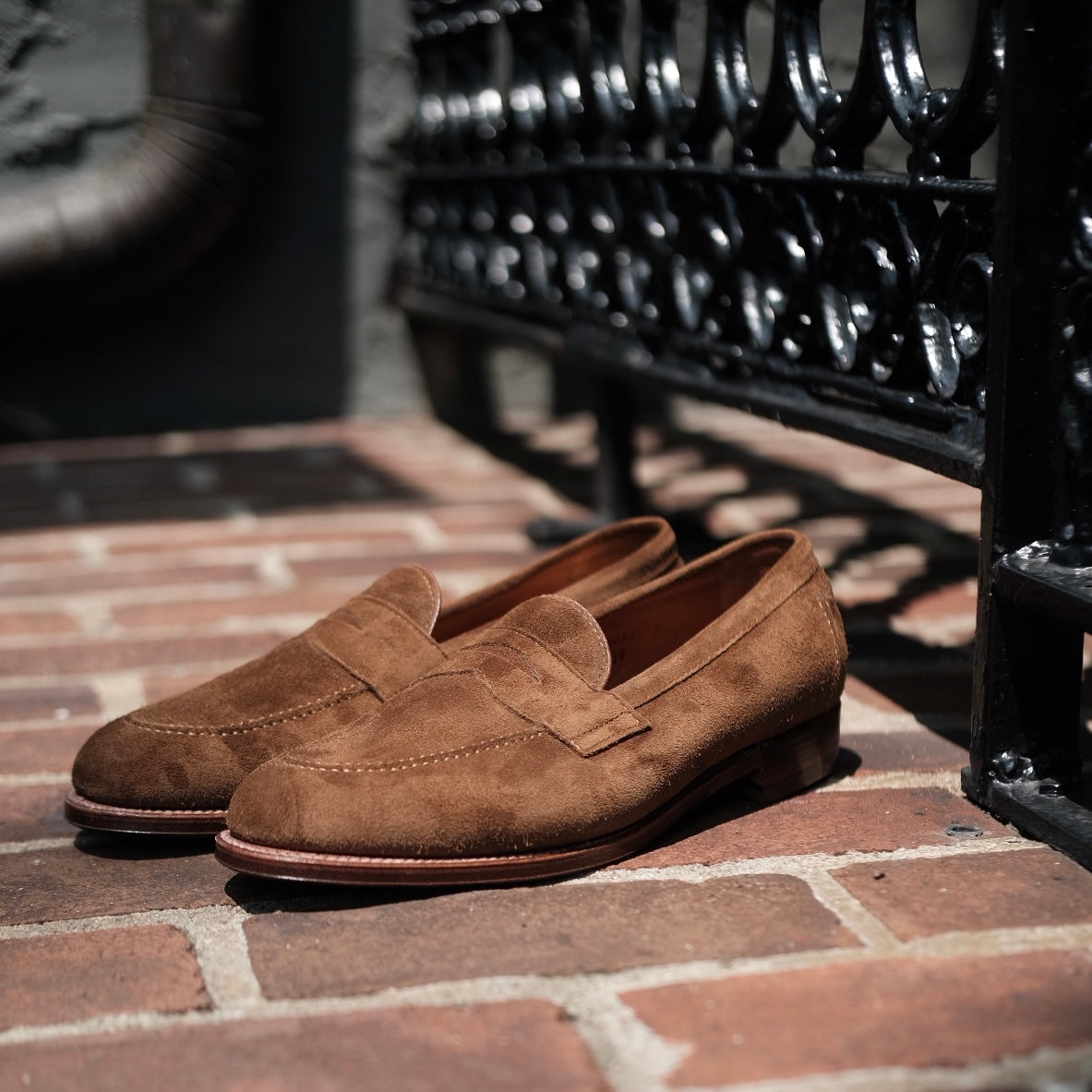 The Alden Madison 9693F Flex Penny Loafer in snuff suede rests on sunlit brick pavement beside a decorative black wrought iron fence.
