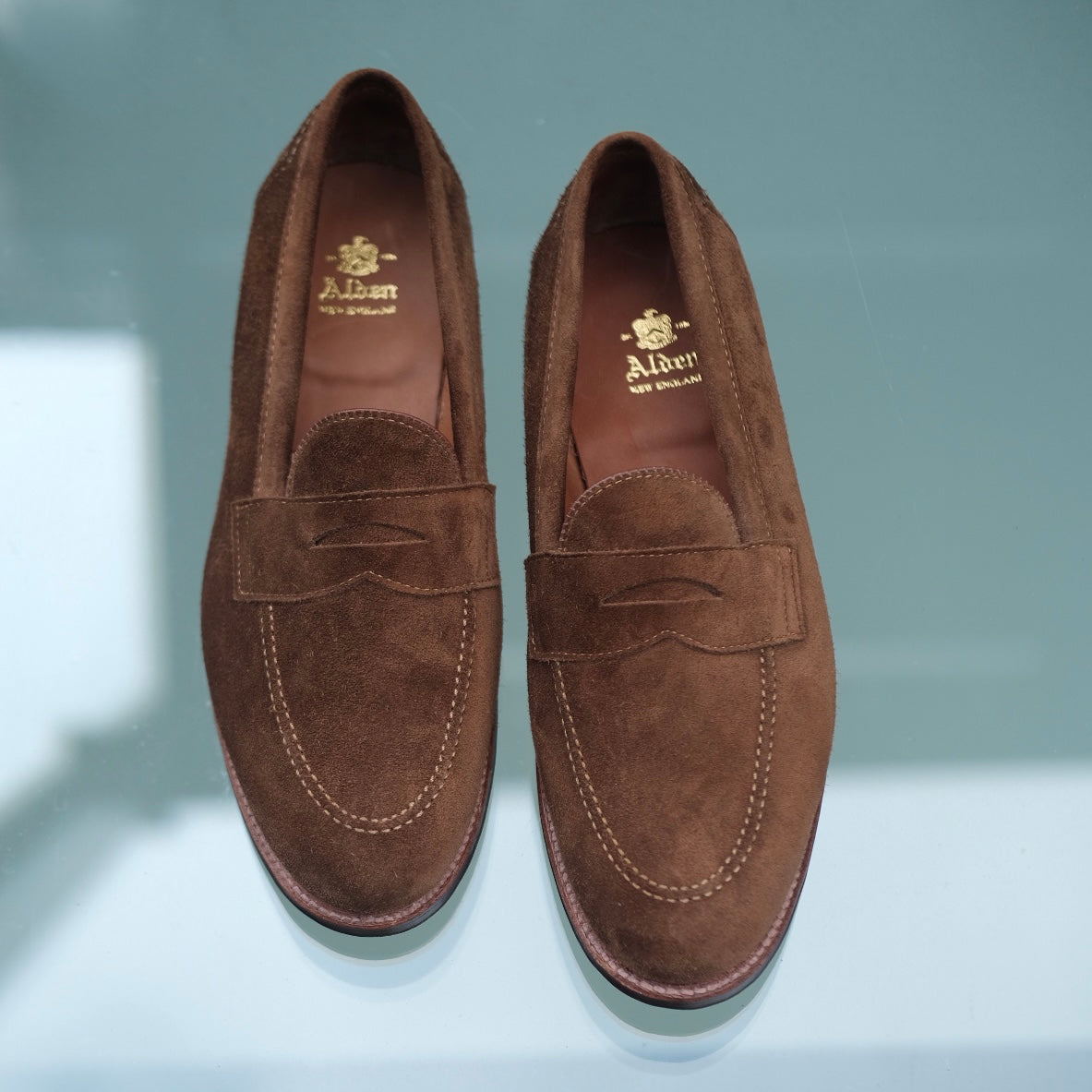 The Alden Madison 9693F Flex Penny Loafers in snuff suede, featuring stitching details and gold branding on the insoles, are displayed side by side on a glass surface.