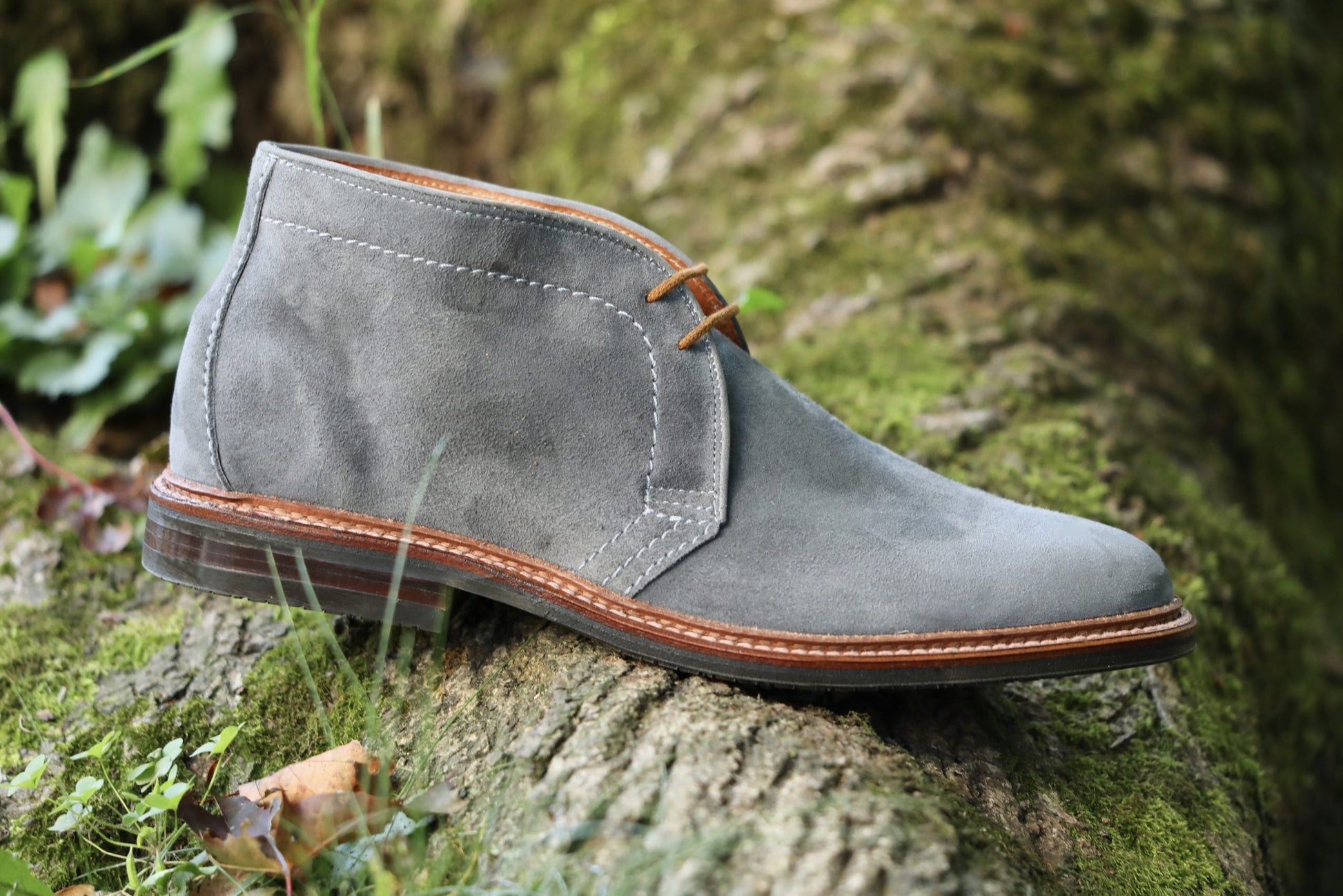 The Alden Madison 1592L Chukka Boot with L3 Sole in Lead Suede, featuring brown laces and stitching, rests on a moss-covered log outdoors amid lush greenery.