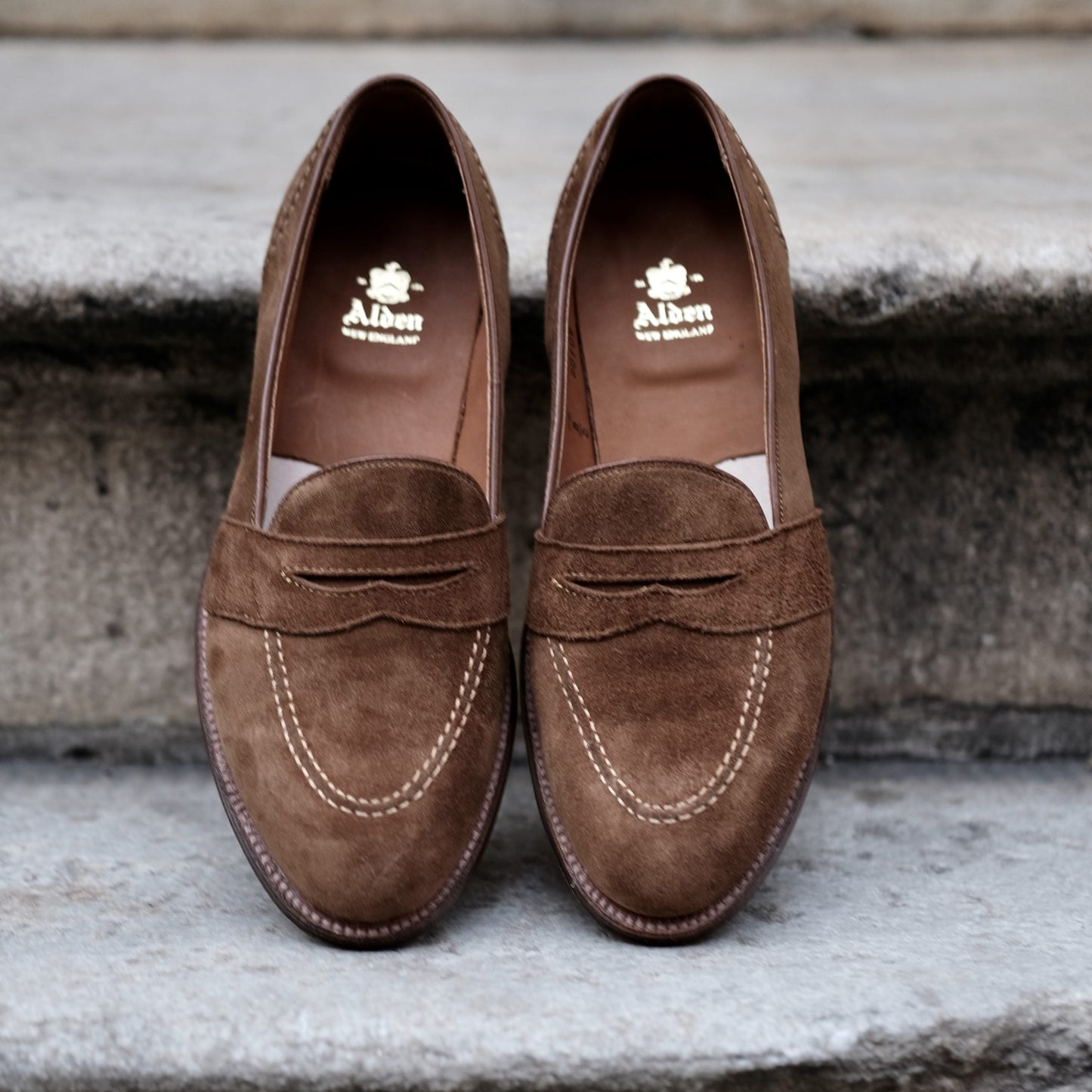A pair of D0109 - Full Strap Loafers in snuff suede by Alden Madison, featuring a leather outsole and white stitching detail, sits on a concrete step. The interior of the left shoe shows the brand name Alden.