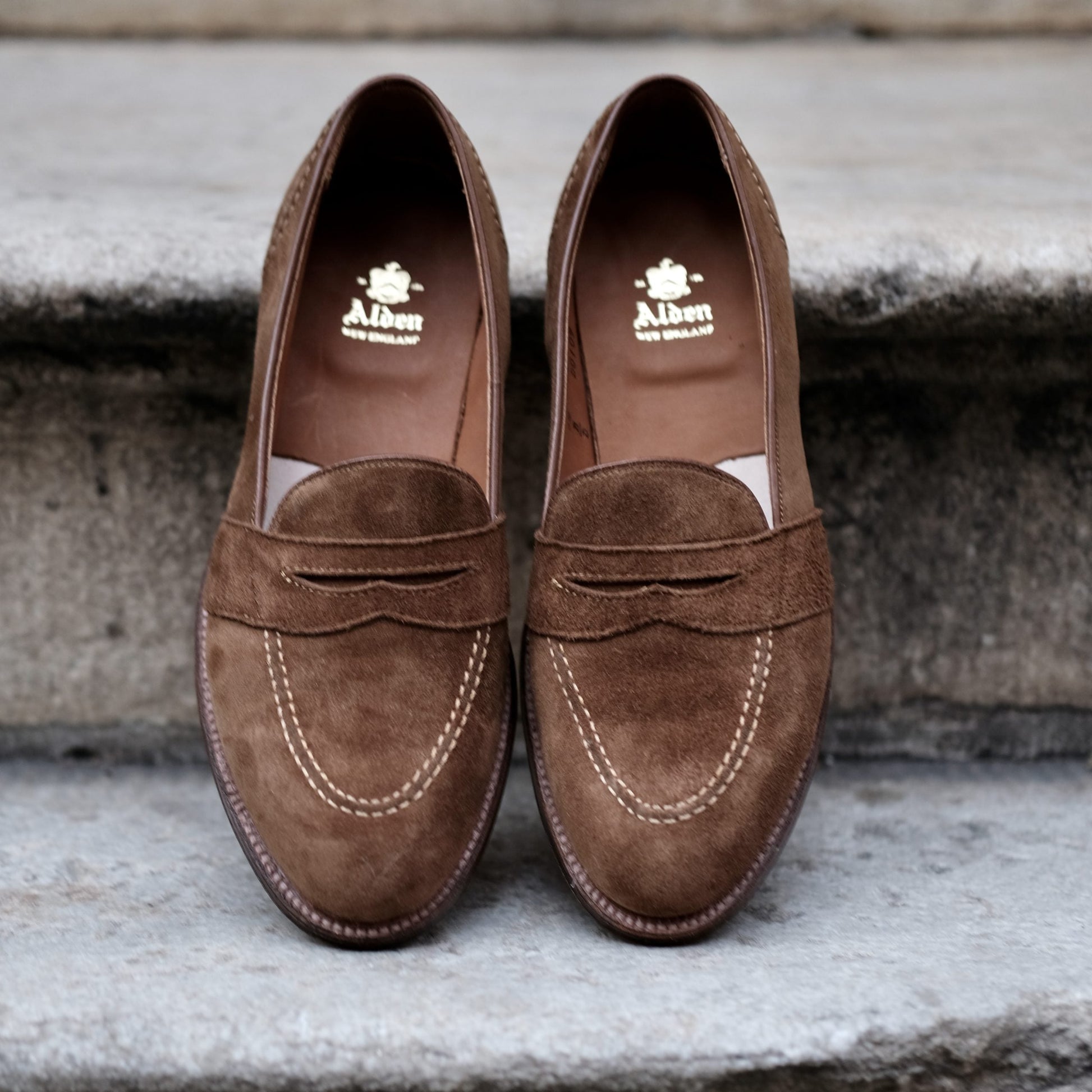 A pair of D0109 - Full Strap Loafers in snuff suede by Alden Madison, featuring a leather outsole and white stitching detail, sits on a concrete step. The interior of the left shoe shows the brand name Alden.