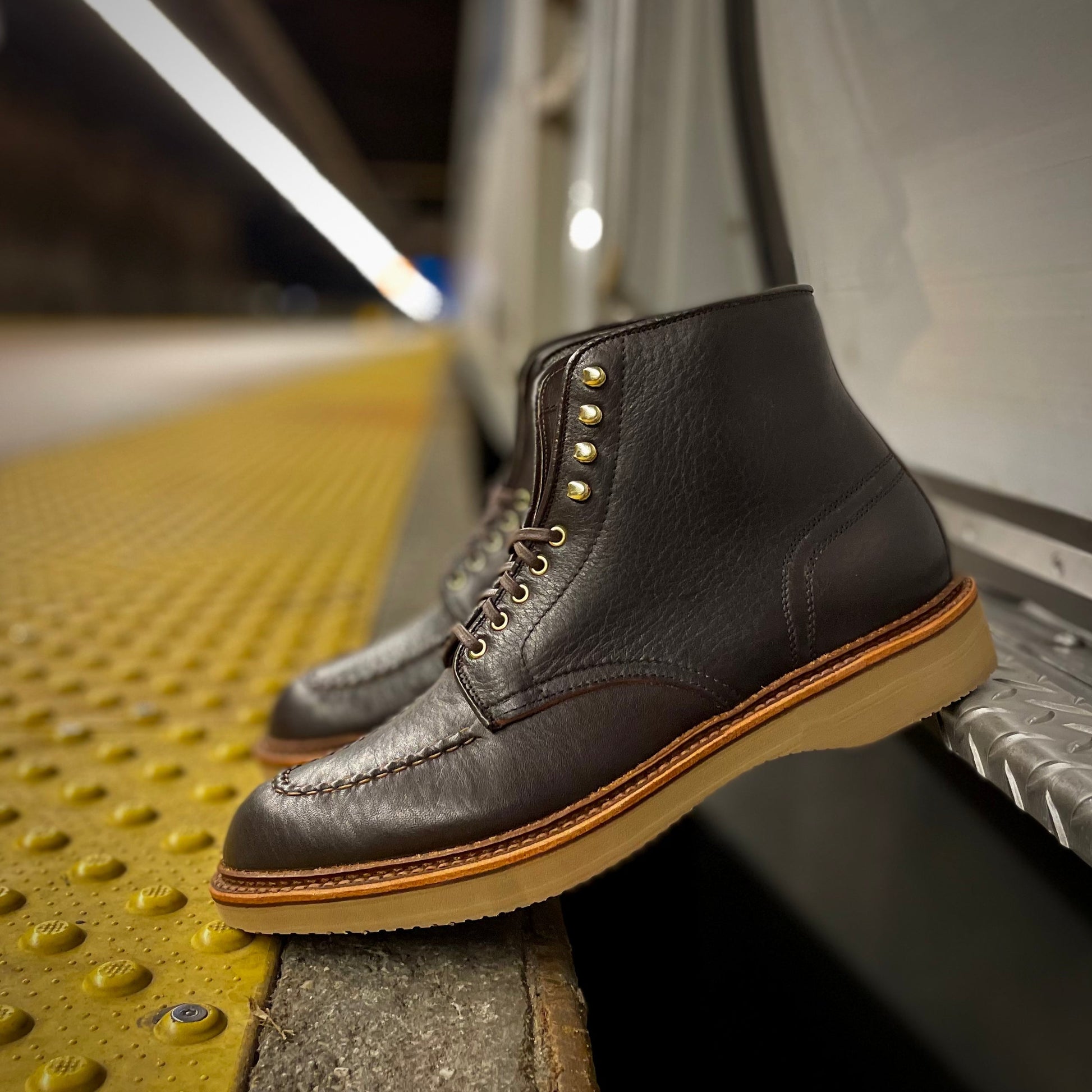 The D1956H - Dirty Bones Indy Boot 2.0 in Arabica Lux by Alden, crafted on the Trubalance Last, is showcased on a subway platform edge with brass eyelets and tan soles, against yellow tactile paving, highlighting its craftsmanship through a shallow depth of field.