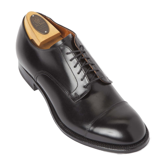 The Alden Madison 558 is a sleek black calfskin dress shoe with a modified cap toe and Single Oak Leather Sole. Black laces enhance its elegance, while a wooden shoe tree maintains its shape, all set against a plain white background.