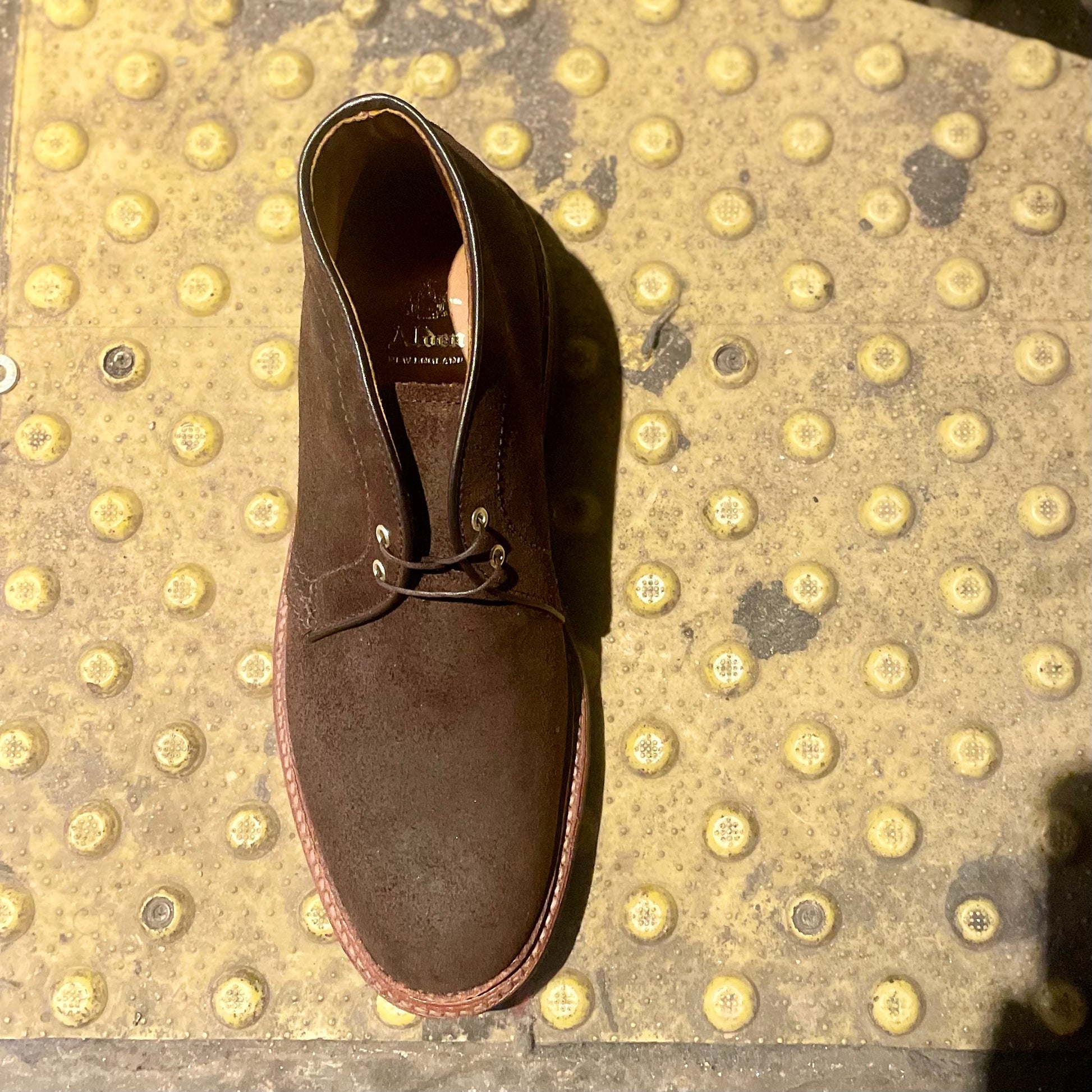 The Alden Madison D1717C Catskills Chukka Boot in Tobacco Reverse Chamois, featuring a Commando sole, is displayed on a yellow textured surface with raised bumps typically found at train platform edges for tactile warning.