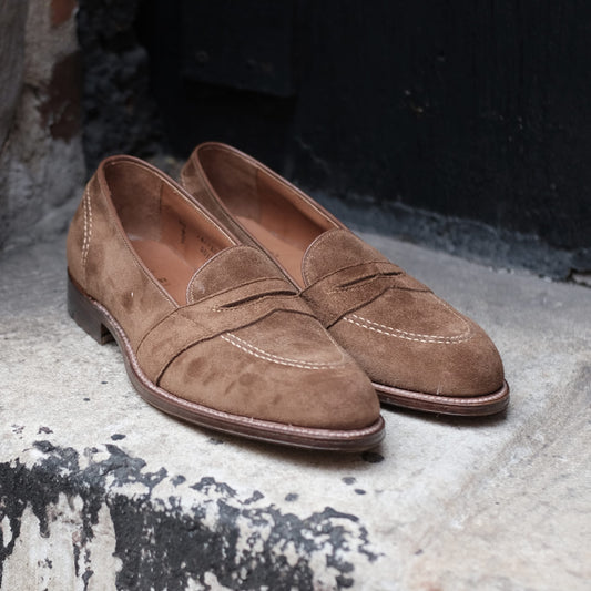 The D0109 - Full Strap Loafer in Snuff Suede from Alden Madison rests on a concrete step. Featuring a penny slot on the vamp, visible stitch pattern, and dark leather outsole, they exude classic elegance against a dark textured wall.