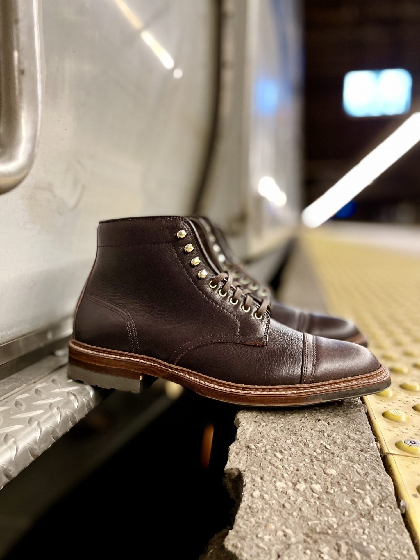 A pair of Alden Madison D1808HC - Stitchup 2/2 Boots in Arabica Lux7 calfskin, featuring metal eyelets, contrast stitching, and a Commando Sole, rest on concrete beside a metallic wall, evoking an industrial or train station vibe.