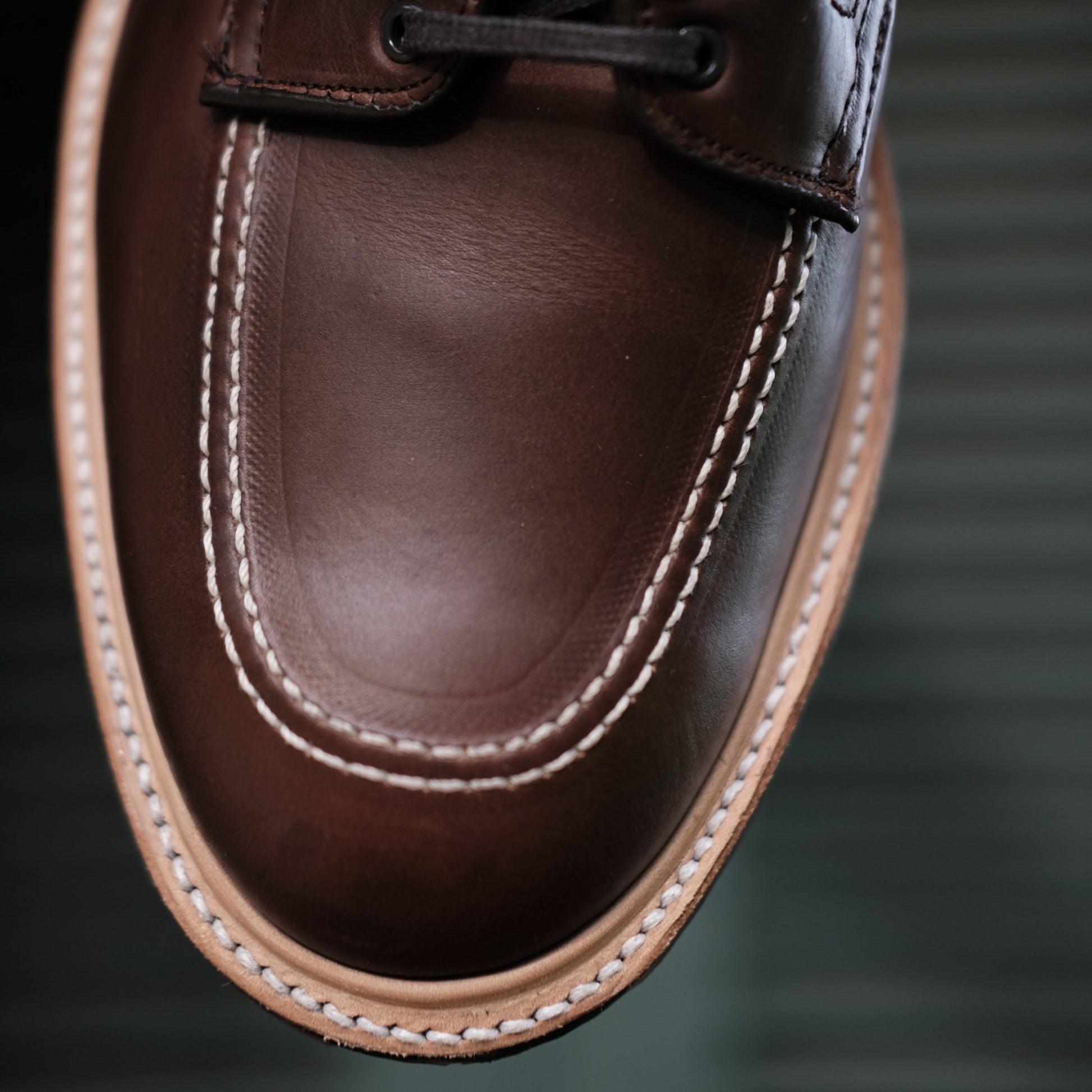 Close-up of the Alden Madison 403C - Indy Boot w Commando Sole in CXL, highlighting its detailed stitching around the toe on rich brown Chromexcel leather. The polished texture and light tan sole edge crafted on a Truebalance Last stand out against a blurred horizontal line background.