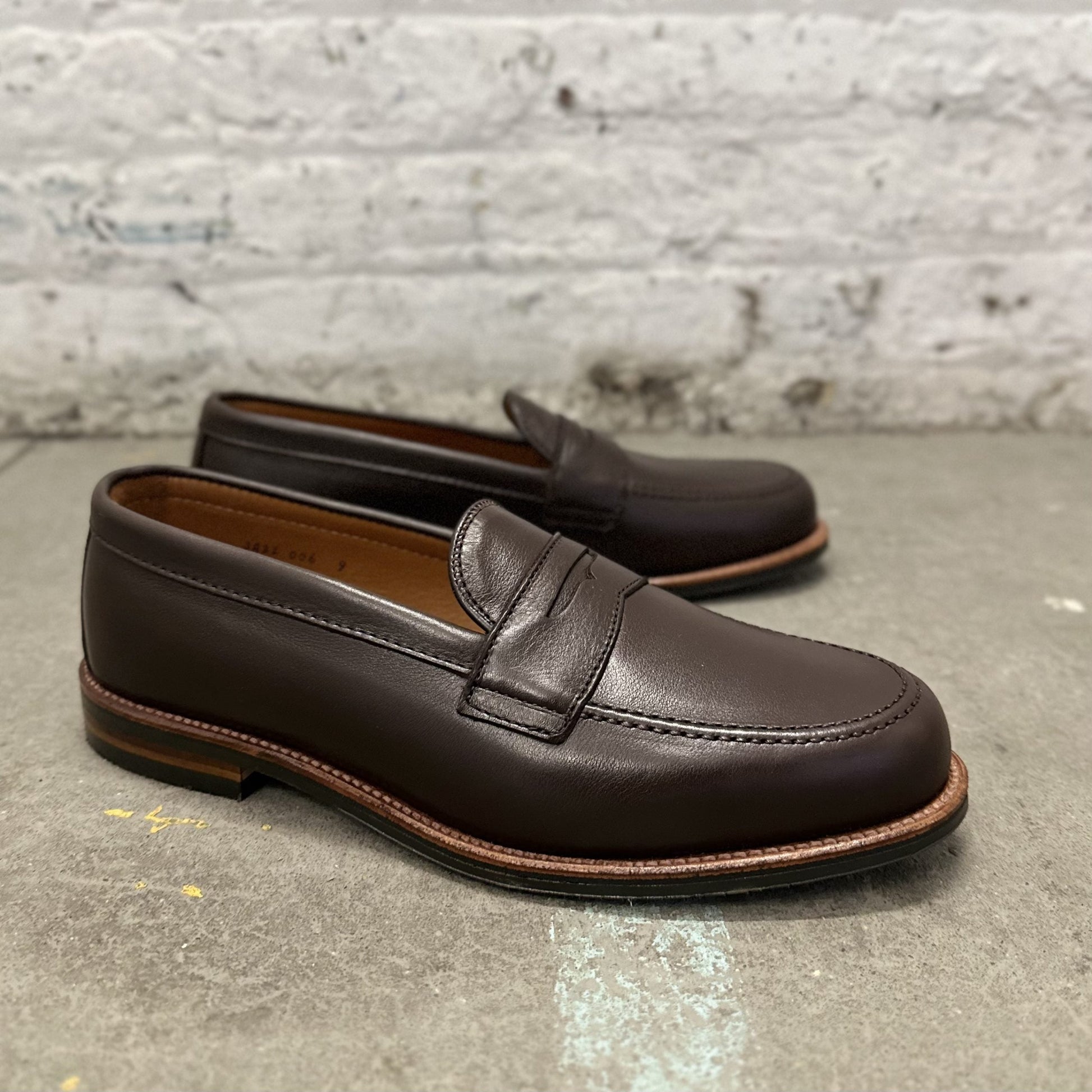 A pair of Alden Madisons D2203L - Penny Loafer in Brown Lady Calf, featuring an antique flat welt and stitching detail, rests on a gray concrete surface against a textured white brick wall, highlighting their classic design.