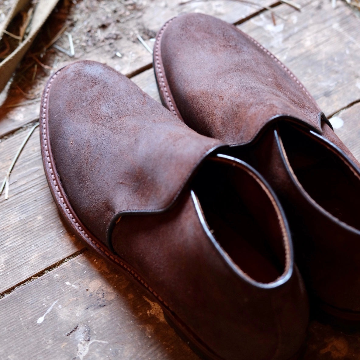 The Alden Madison D8704C Folsom Slip On Chukka in Tobacco Reverse Chamois, featuring commando rubber soles, rests on a rustic wooden floor with dust and straw scattered around, evoking a laid-back, broken-in vibe.