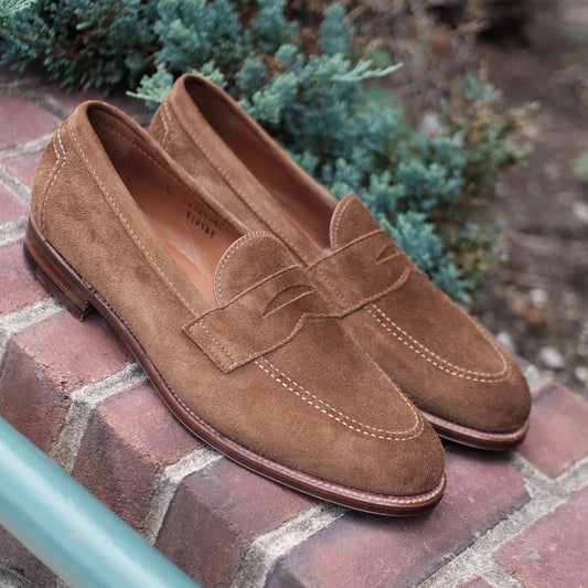 The Alden Madison 9693F Flex Penny Loafer in snuff suede, featuring stitching detail and a flex leather sole, sits on a brick ledge outdoors with green foliage in the background.