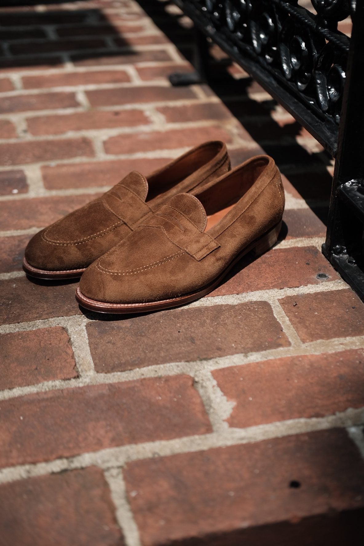 A pair of Alden Madison 9693F - Flex Penny Loafer in Snuff Suede with Flex Leather Soles rests on a brick sidewalk in sunlight, next to a black wrought-iron fence.