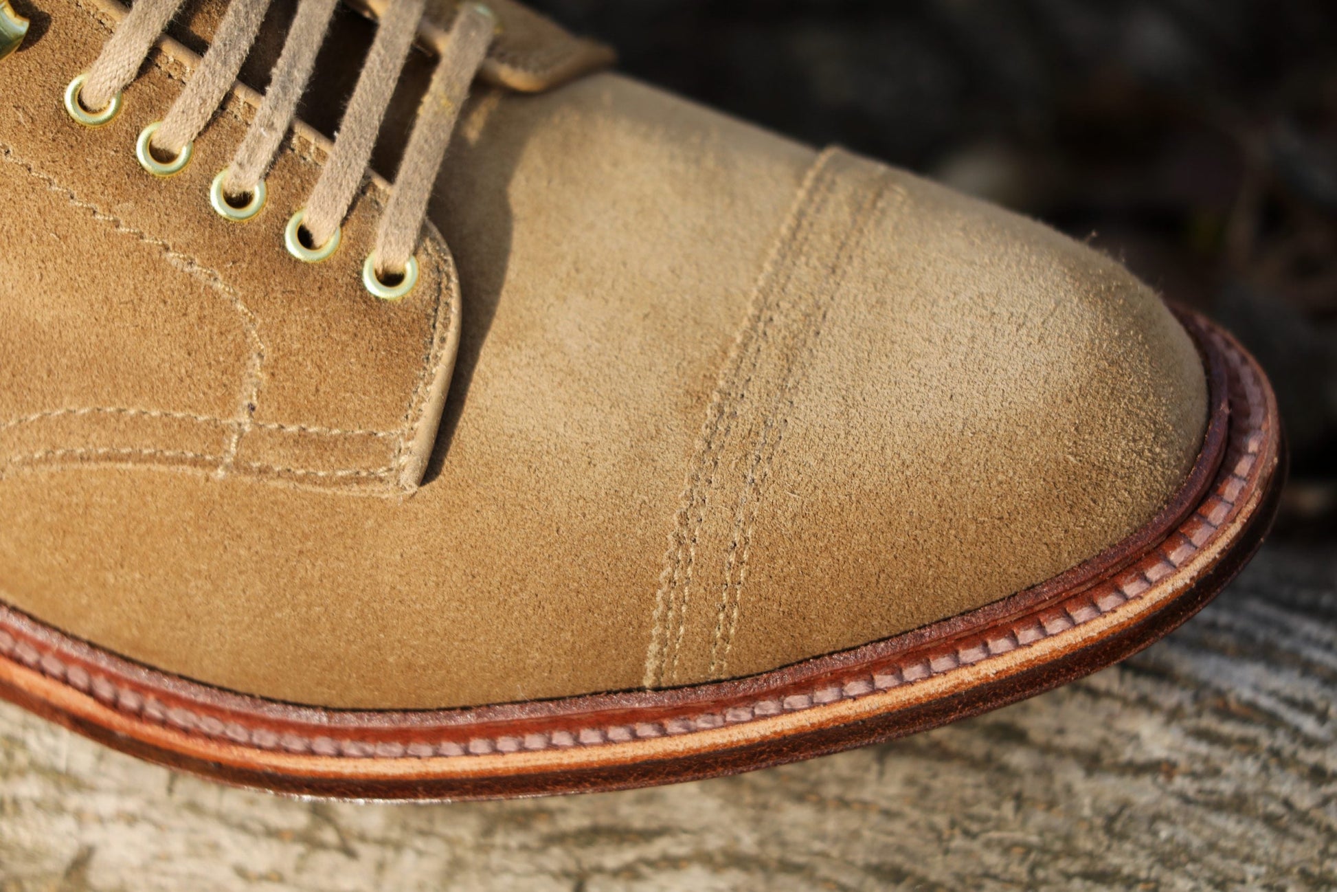Close-up of the Alden Madison D2685H - 2/2 Cap Toe Boot in Tan Suede with detailed toe cap stitching, brass eyelets, and a welted sole, elegantly resting on a textured surface.