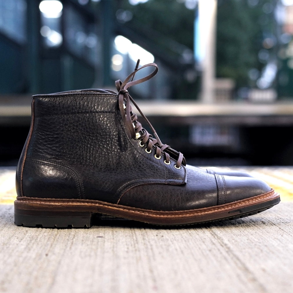A close-up side view of the Alden Madison D1808HC - Stitchup 2/2 Boot in Arabica Lux7 calfskin shows its dark brown grained leather, metal eyelets, tan welted sole, and lace-up design, set on concrete with a blurred outdoor background.