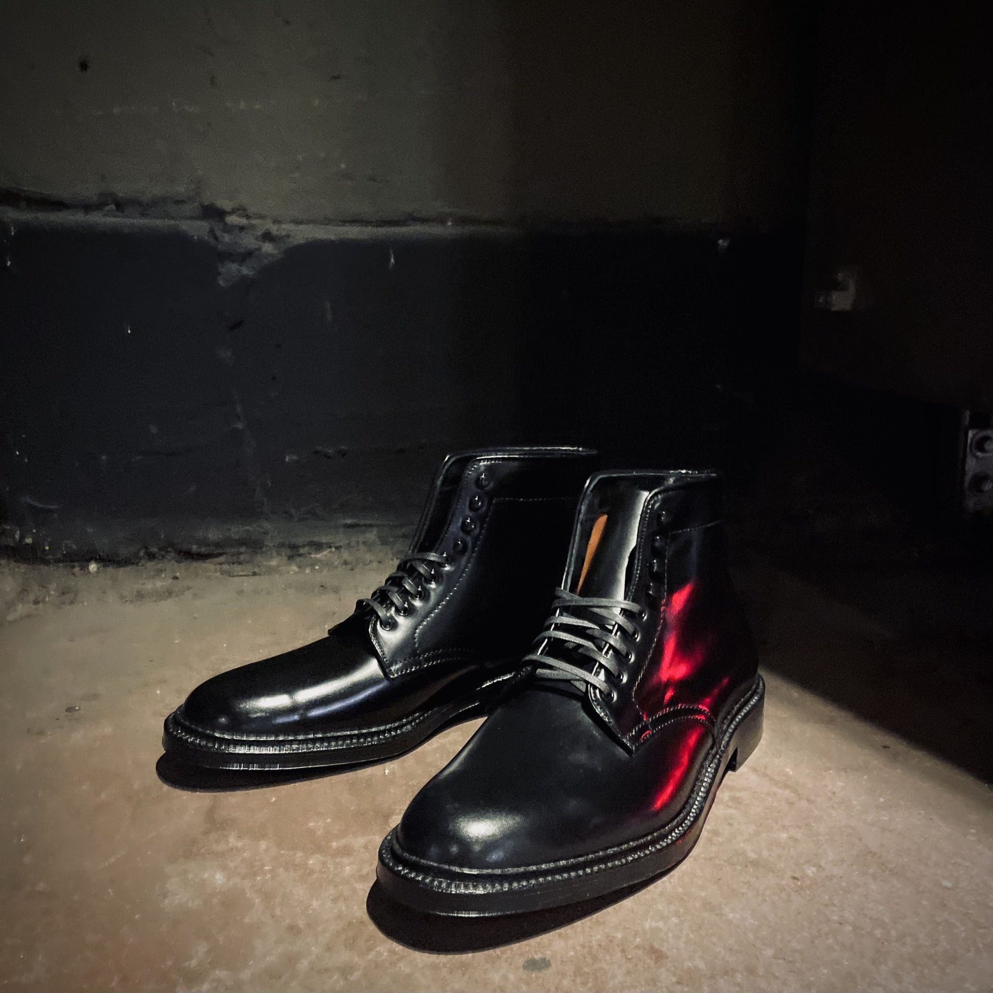 The D6842H Director Boot in Black Shell Cordovan from Alden Madison rests on a dimly lit concrete floor, casting shadows against a dark wall.