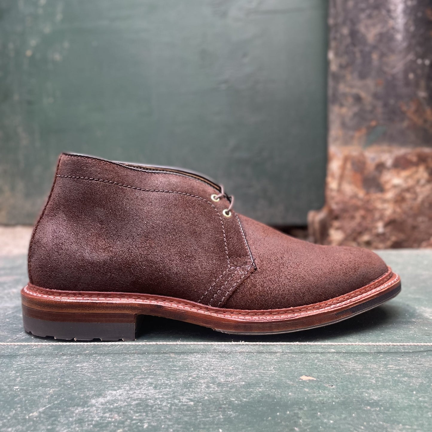 The Alden Madison D1717C - Catskills Chukka Boot in Tobacco Reverse Chamois features dark laces and a wooden sole, displayed on a green, worn wooden surface with a weathered wall in the background.