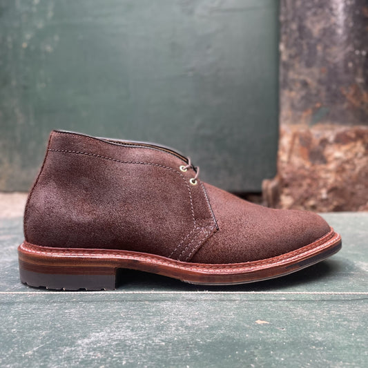 The Alden Madison D1717C - Catskills Chukka Boot in Tobacco Reverse Chamois features dark laces and a wooden sole, displayed on a green, worn wooden surface with a weathered wall in the background.