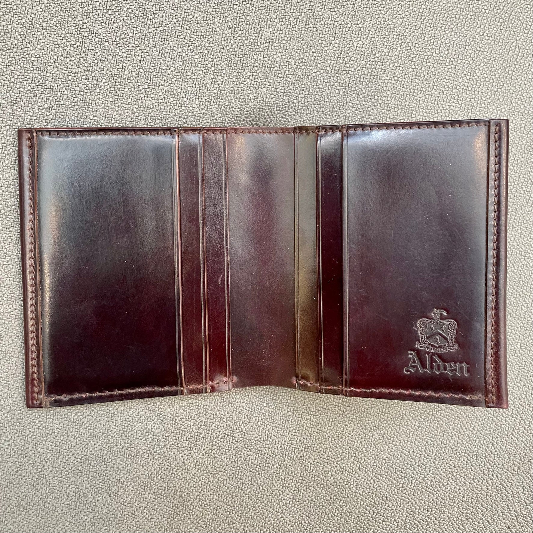The Alden Madison LG 7008 - Alden Color 8 Shell Cordovan Wallet, a three-section leather wallet with multiple card slots and an embossed logo, is open and empty on a textured gray surface.