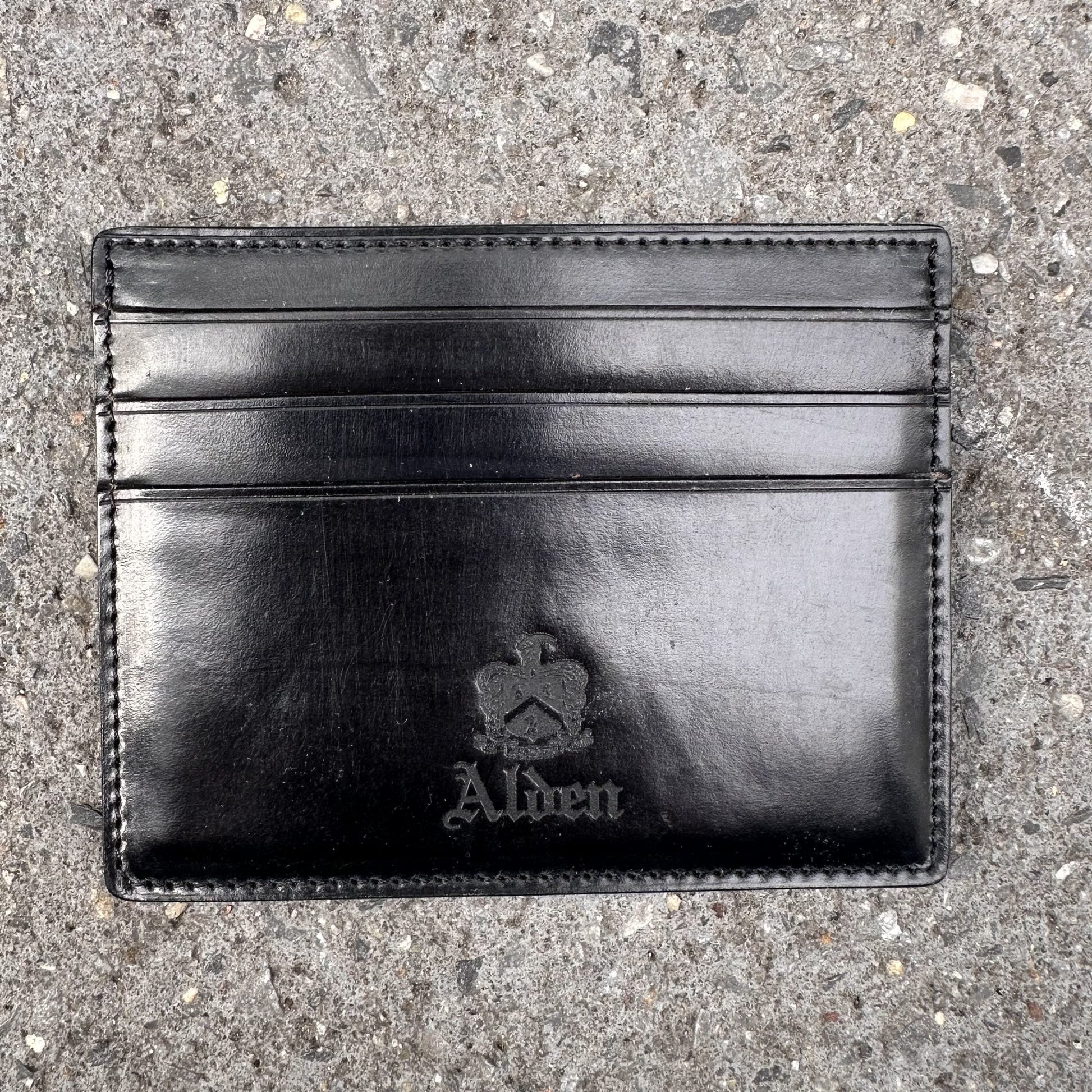 The Alden Madison LG 6009 is a black leather Shell Cordovan cardholder embossed with Alden and a crown logo, featuring six pockets for essentials, elegantly displayed on textured stone.