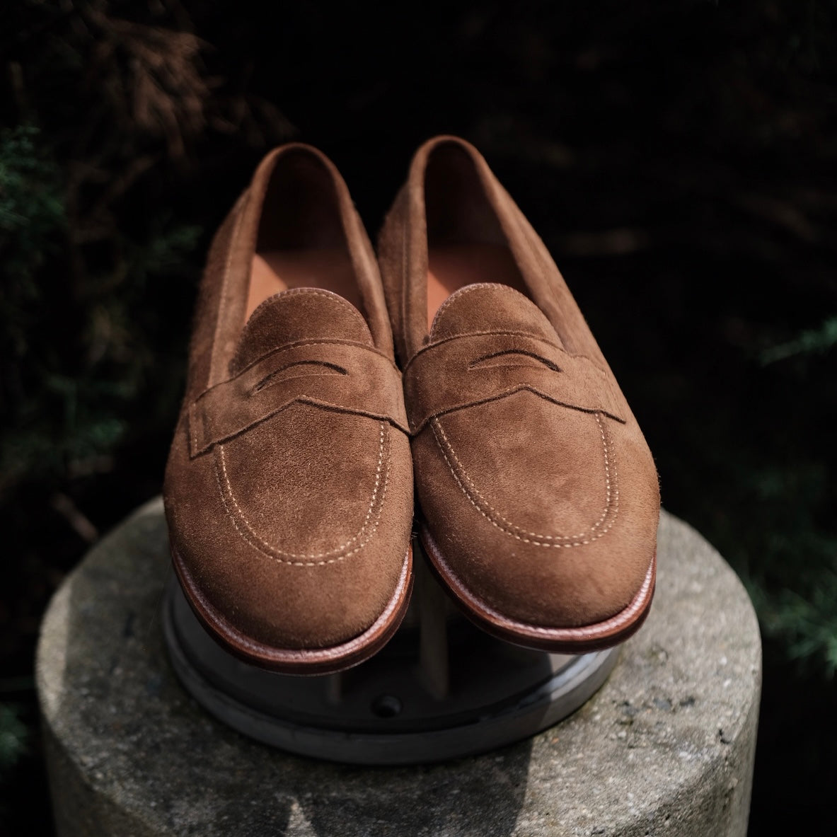 The Alden Madison 9693F - Flex Penny Loafer in snuff suede, featuring stitched detailing and a flex leather sole, is shown outdoors on a round concrete pedestal with blurred greenery in the background.
