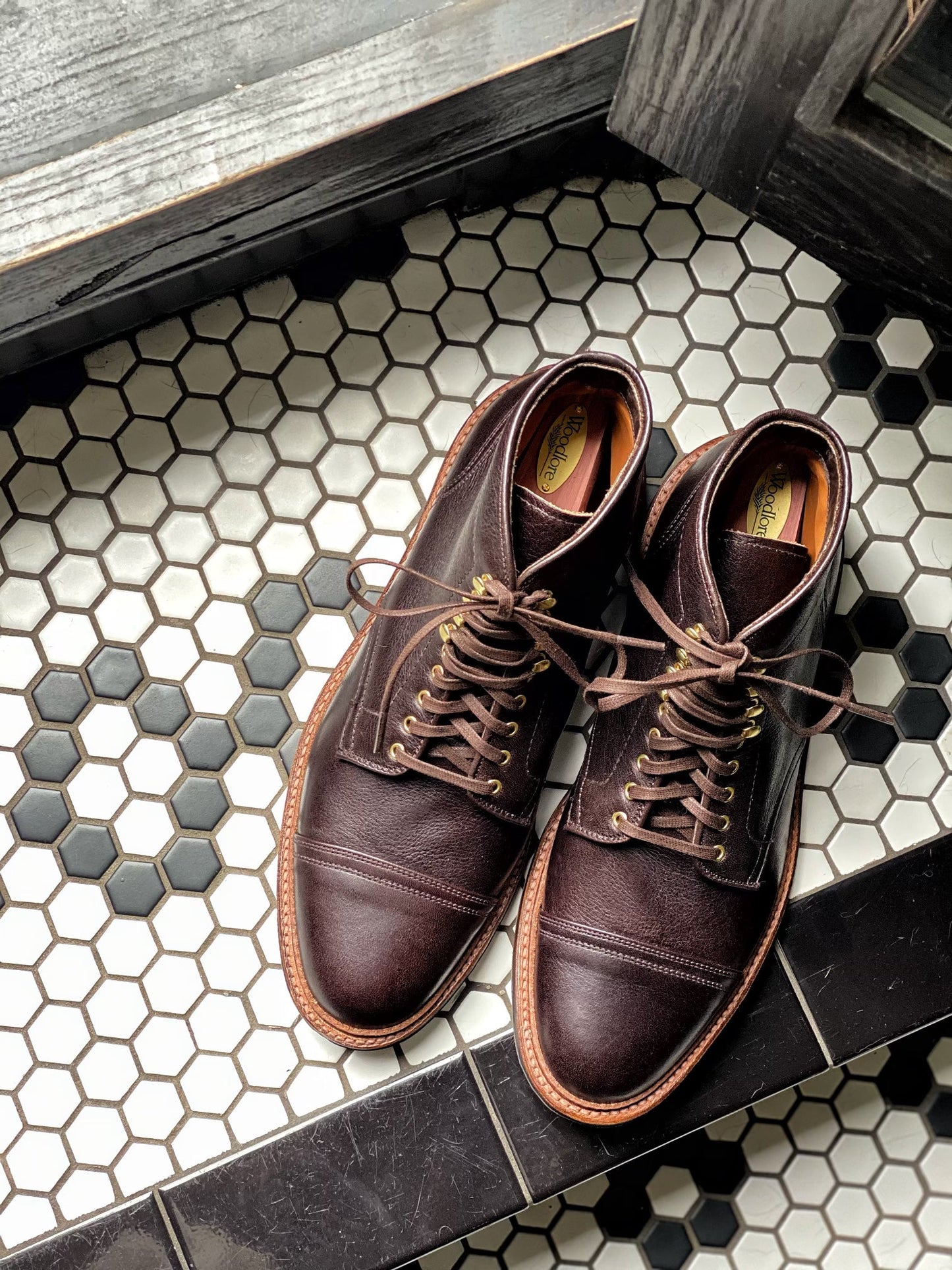 A pair of Alden Madison D1808HC - Stitchup 2/2 Boots in Arabica Lux7 with sturdy Commando soles rests on black and white hexagonal tiles near a window or doorway.