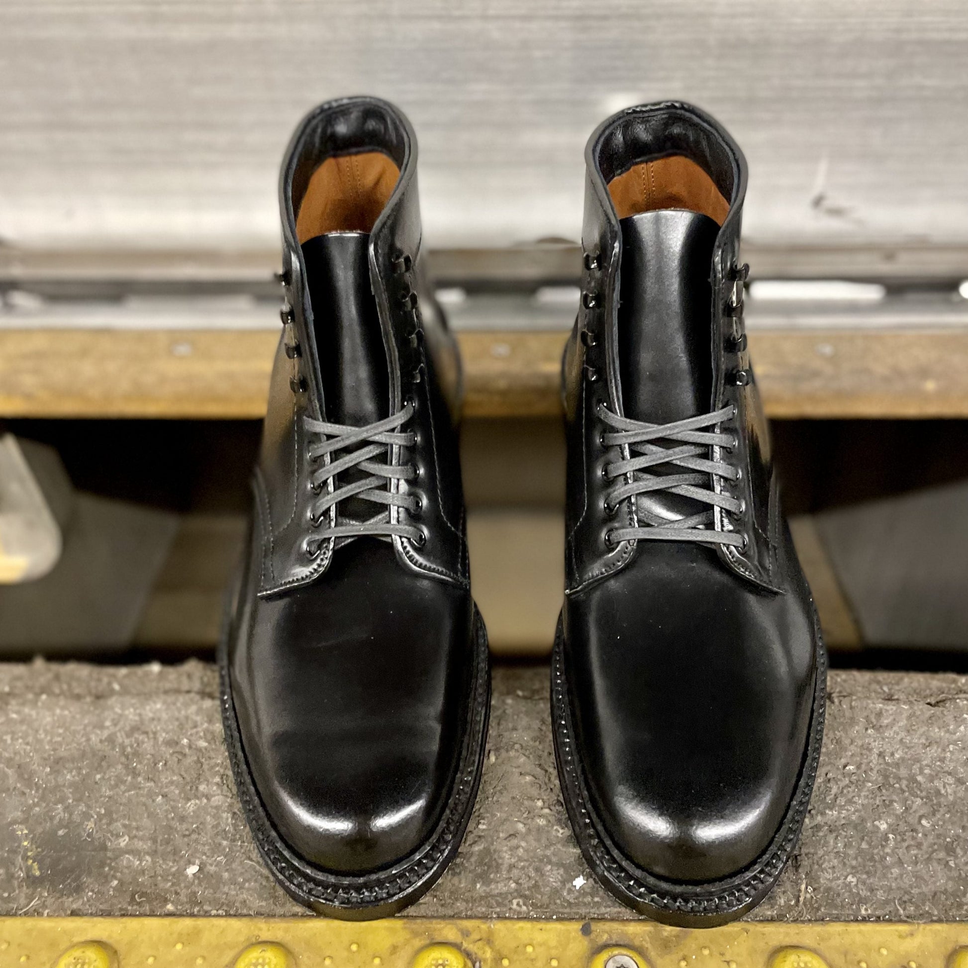 The Alden Madison Director boots, model D6842H in Black Shell Cordovan, feature sleek shiny leather and gray laces with speed hooks. These stylish boots, with a double oak leather sole, are placed together on a textured surface near a metallic background.