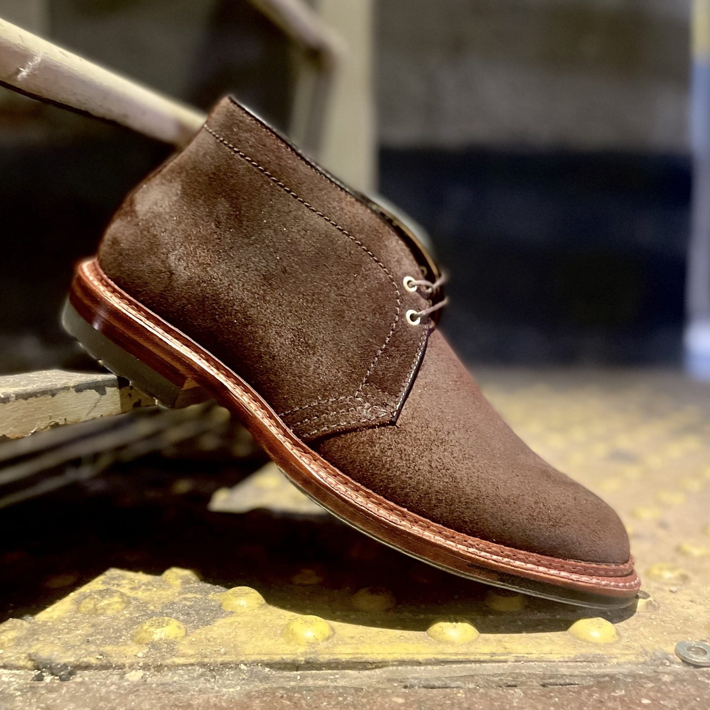 The Alden Madison D1717C Catskills Chukka Boot in Tobacco Reverse Chamois, featuring a Commando sole and metal eyelets, is propped on its heel against a worn staircase, showcasing its textured suede and fine stitching.