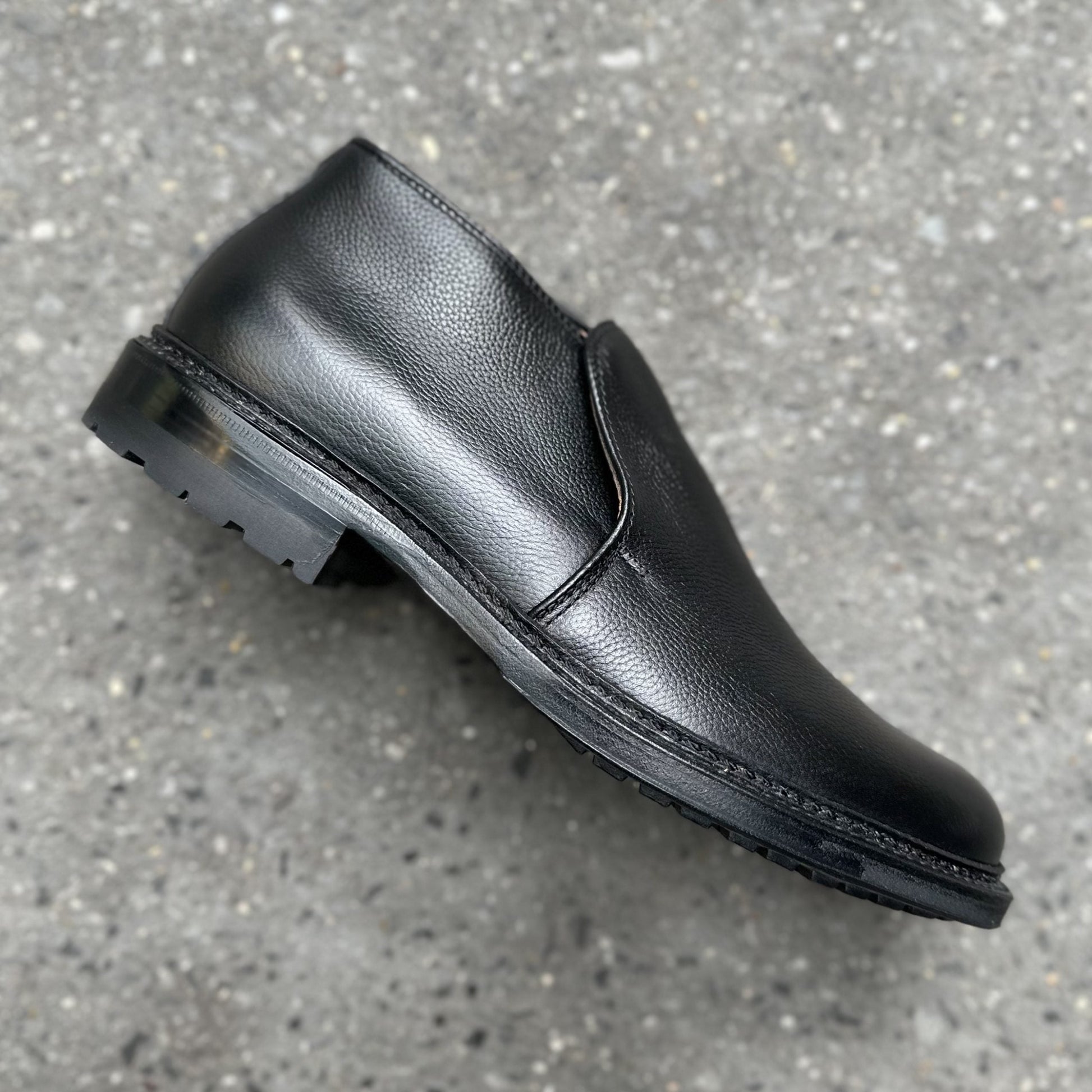 A single Alden Madison D2711C - 750 Chukka Boot in Black Regina Grain, featuring a smooth, pebbled leather texture, rounded toe, and sturdy Commando Rubber Sole, sits on a gray speckled concrete surface.