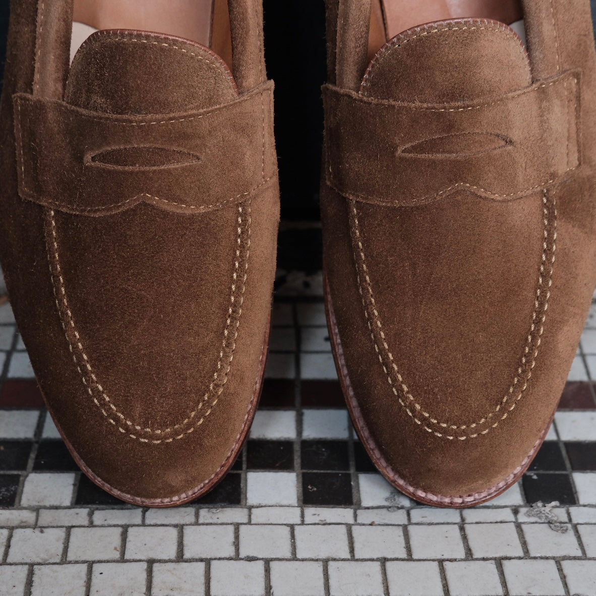 A close-up of the Alden Madison 9693F - Flex Penny Loafer in Snuff Suede, featuring visible stitching and flex leather soles, worn by someone standing on a black, white, and gray tiled floor.