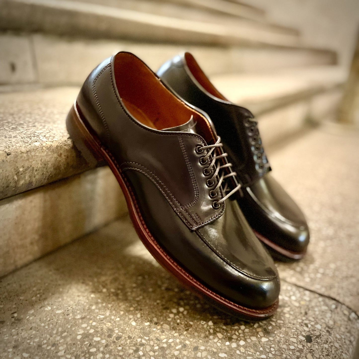 A pair of Alden Madisons D1629C - V Tip Split Toe Blucher in Color 8 Shell shoes, featuring a polished finish and brown soles, sit on a textured stone staircase with neatly tied laces reflecting overhead lighting.