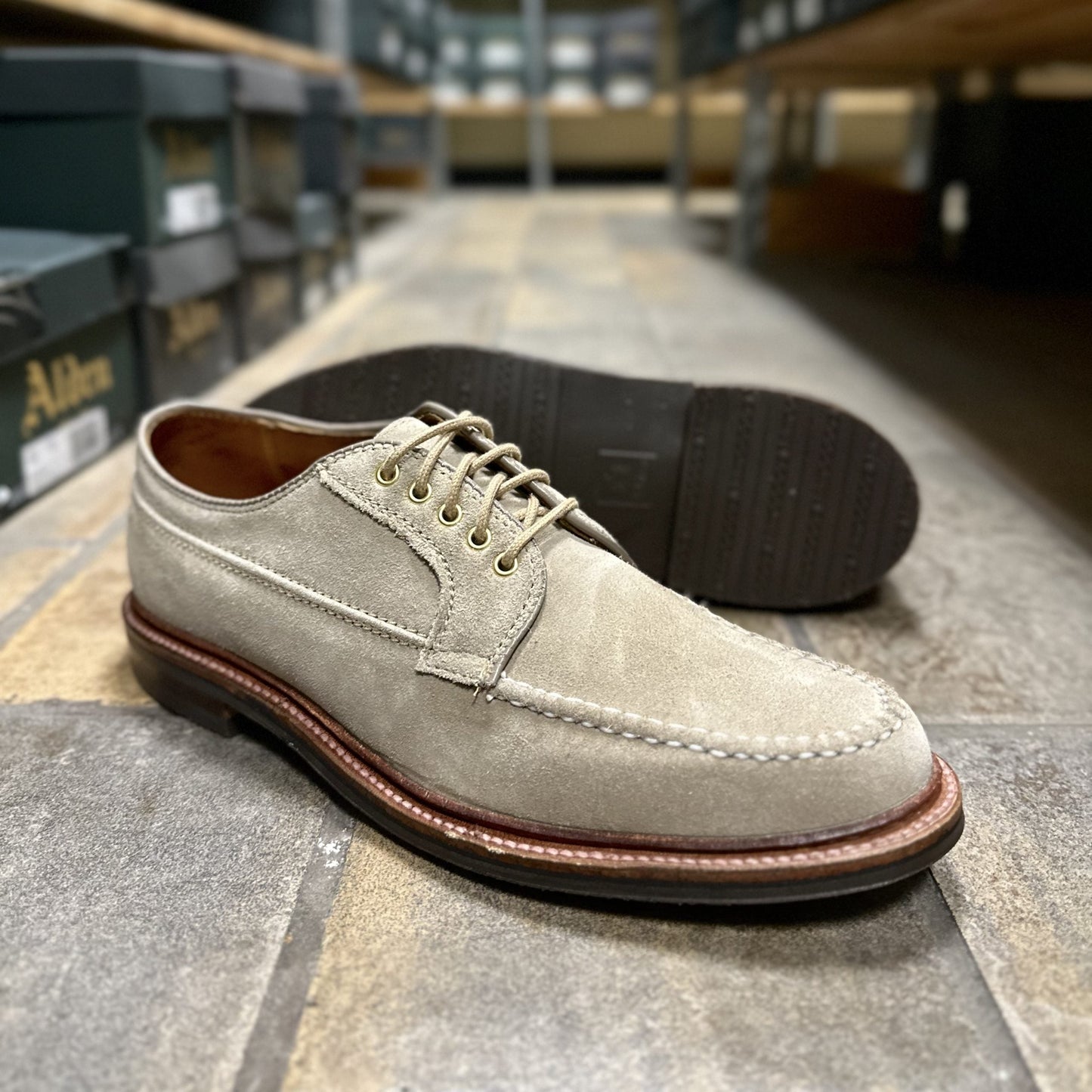The Alden Madison D1634L - Ranger Mocc in Milkshake Suede, featuring brown rubber soles and an Antique 360 Flat Welt, is shown on a tiled floor in a shoe store with shelves and shoe boxes in the background.