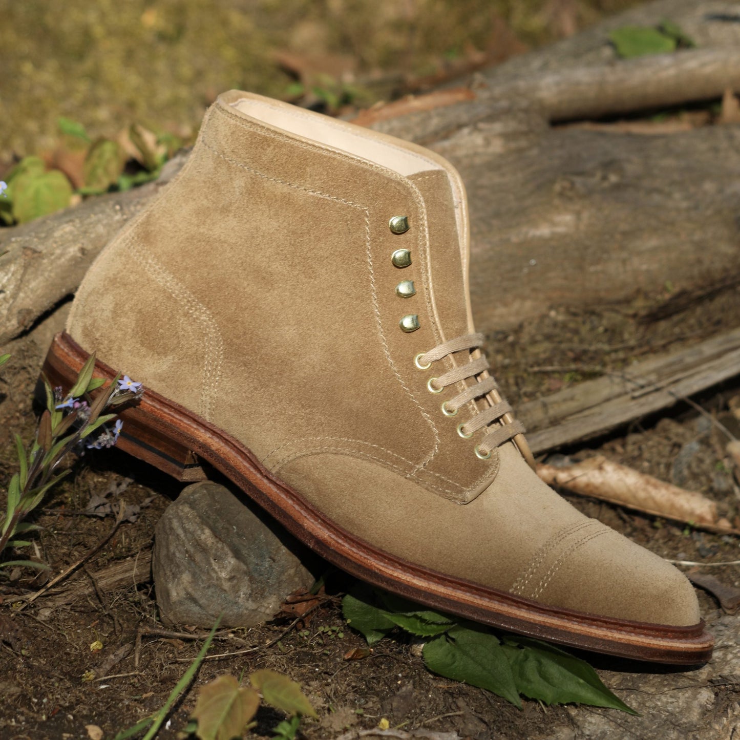 The Alden Madison D2685H - 2/2 Cap Toe Boot in Tan Suede, featuring metallic eyelets, a Barrie last, sturdy wooden sole, and classic cap toe design, rests on its side in a natural outdoor setting among wooden logs and flowers.