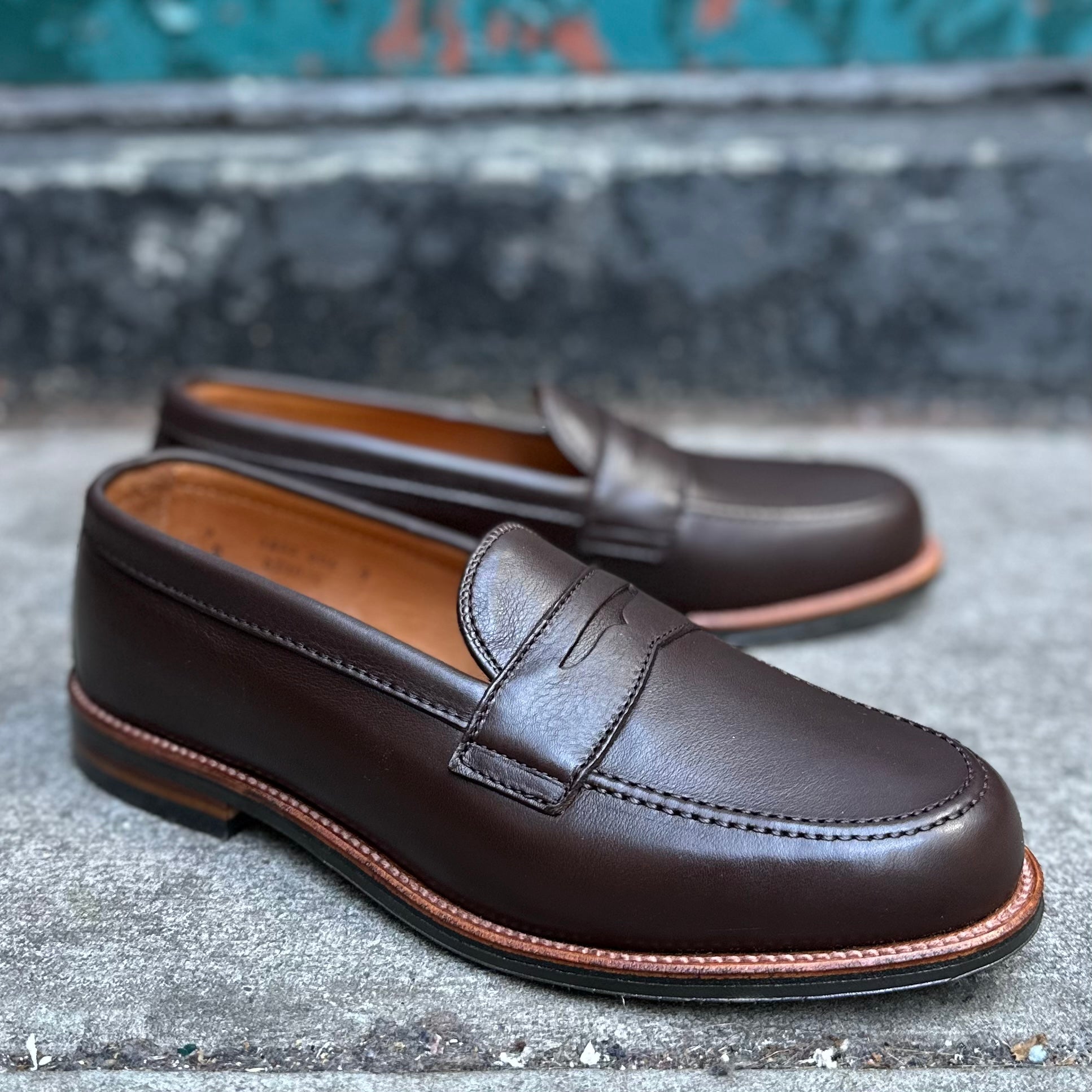 The D2203L Penny Loafer in Brown Lady Calf by Alden Madison features a polished finish and light brown soles. They are positioned on a concrete surface with an Antique Flat Welt design against a worn, teal backdrop.