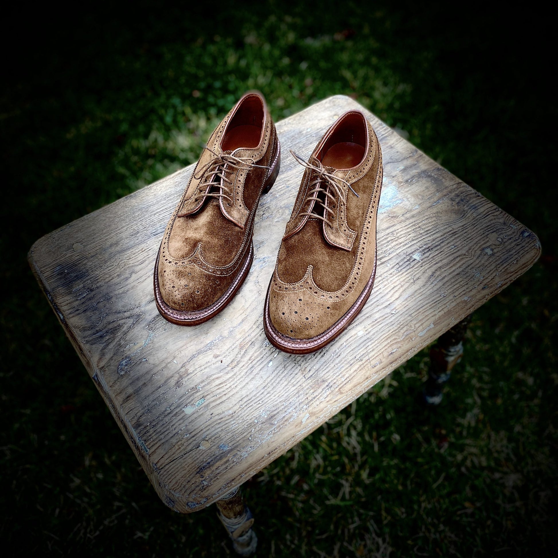 A pair of Alden Madisons 9794 - Long Wing Blucher in Snuff Suede shoes with decorative perforations and a Waterlock leather sole rests on a rustic wooden stool in a grassy outdoor setting, showcasing their laced design and slightly worn appearance.