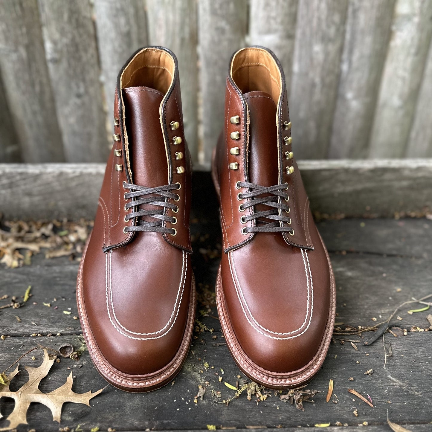 Aldens Madison Indy Boot in Madison Brown Calf, D2940H, is crafted from premium calfskin with white stitching and tan laces. Elegantly resting on a wooden surface, an oak leaf partially visible nearby complements its design.