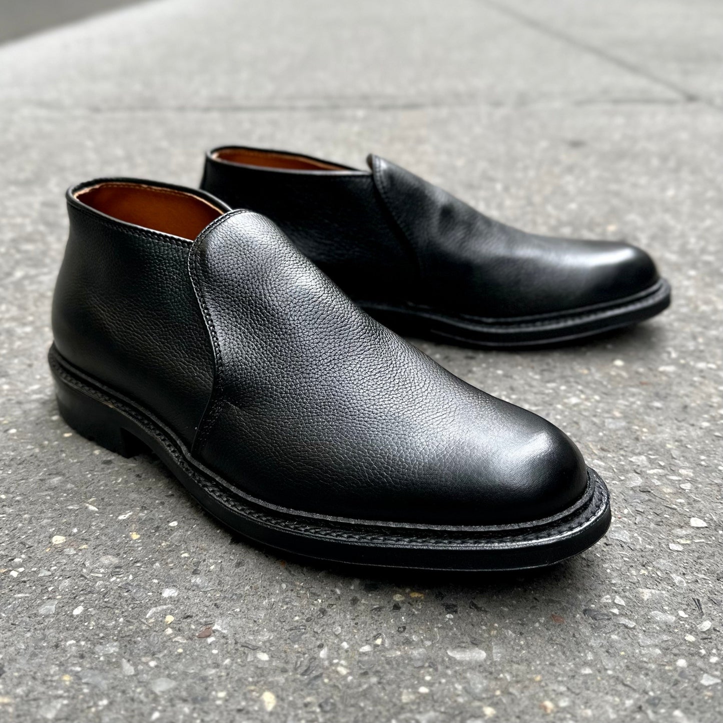 The Alden Madison D2711C - 750 Chukka Boot in Black Regina Grain, featuring rounded toes, visible stitching, and a sturdy Commando sole, is showcased on a gray textured sidewalk.