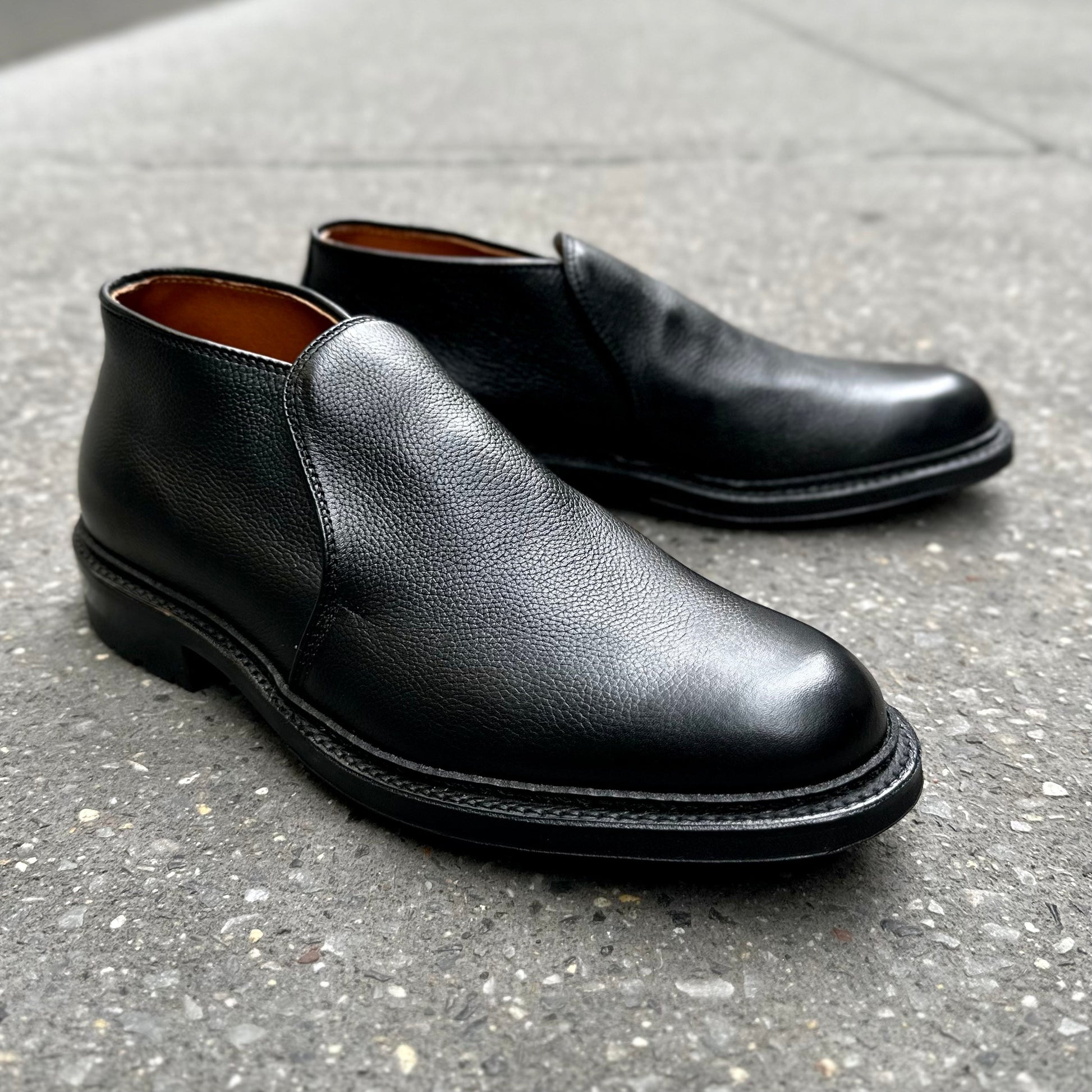 The Alden Madison D2711C - 750 Chukka Boot in Black Regina Grain, featuring rounded toes, visible stitching, and a sturdy Commando sole, is showcased on a gray textured sidewalk.