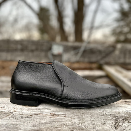 The Alden Madison D2711C - 750 Chukka Boot in Black Regina Grain with a Commando Rubber Sole rests on a weathered wooden surface outdoors, framed by blurred trees and a gray sky.