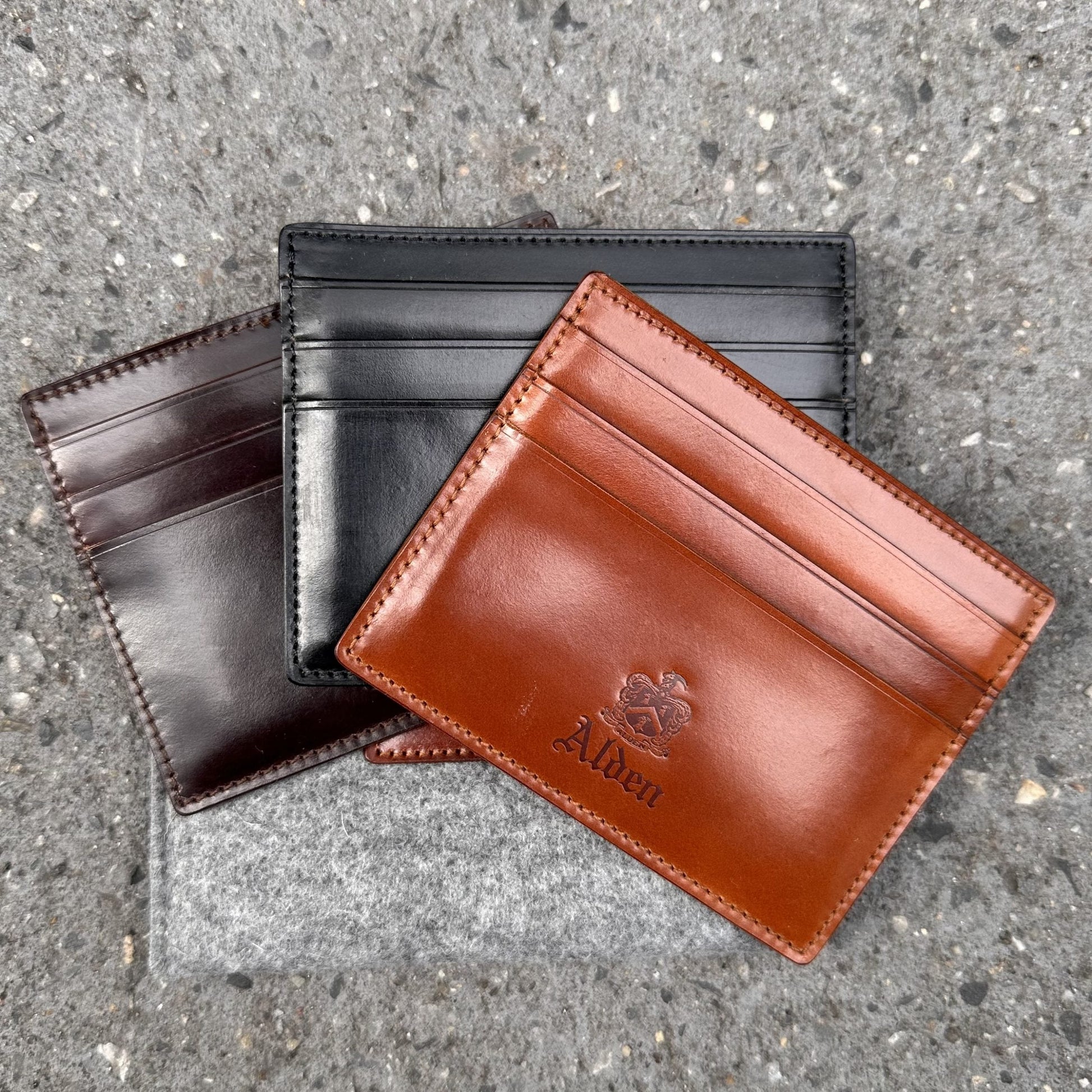 Three leather cardholders—one black, one dark brown, and one tan—lie on a gray textured surface. The tan cardholder is the Alden Madison LG 6008 - Alden Color 8 Shell Credit Card Holder with an embossed logo and features 6 pockets for ample storage.