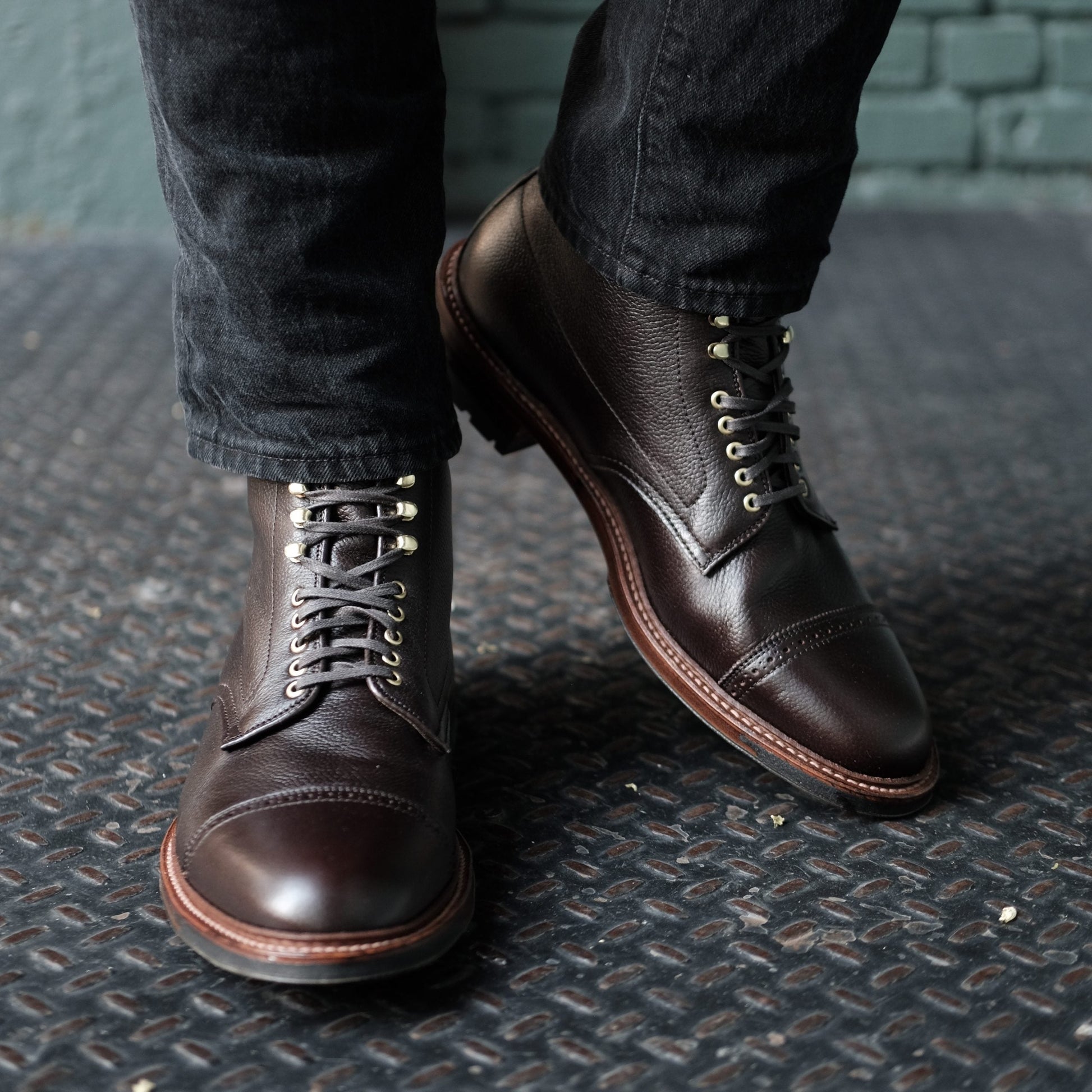 Wearing Alden Madison’s D9819HC Jumper Boot in Regina Brown Grain, a person pairs dark leather lace-up boots with black pants, standing on a textured metal surface against a greenish-gray brick wall.