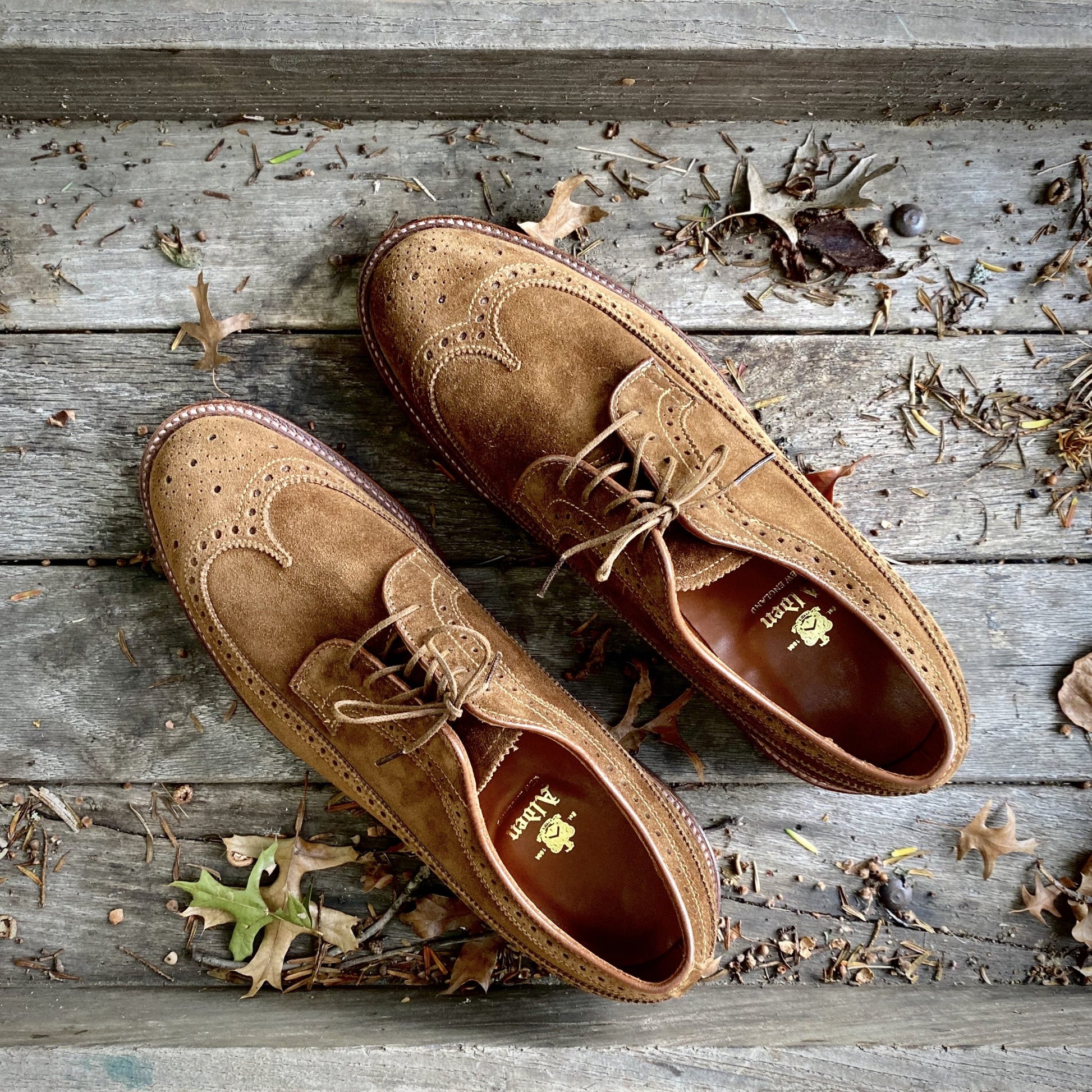 The Alden Madison 9794 Long Wing Blucher in snuff suede sits on a weathered wooden surface, surrounded by scattered dry leaves and twigs for an autumnal vibe. Detailed stitching and laces enhance its design, while the Waterlock leather sole ensures durability.