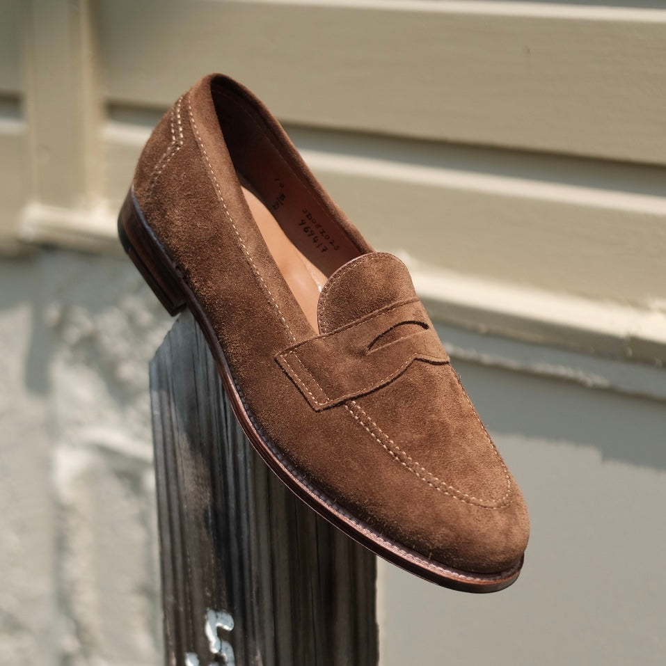 The Alden Madison 9693F - Flex Penny Loafer in snuff suede, featuring a classic penny strap detail, is showcased atop a wooden post before a cream-colored wall.