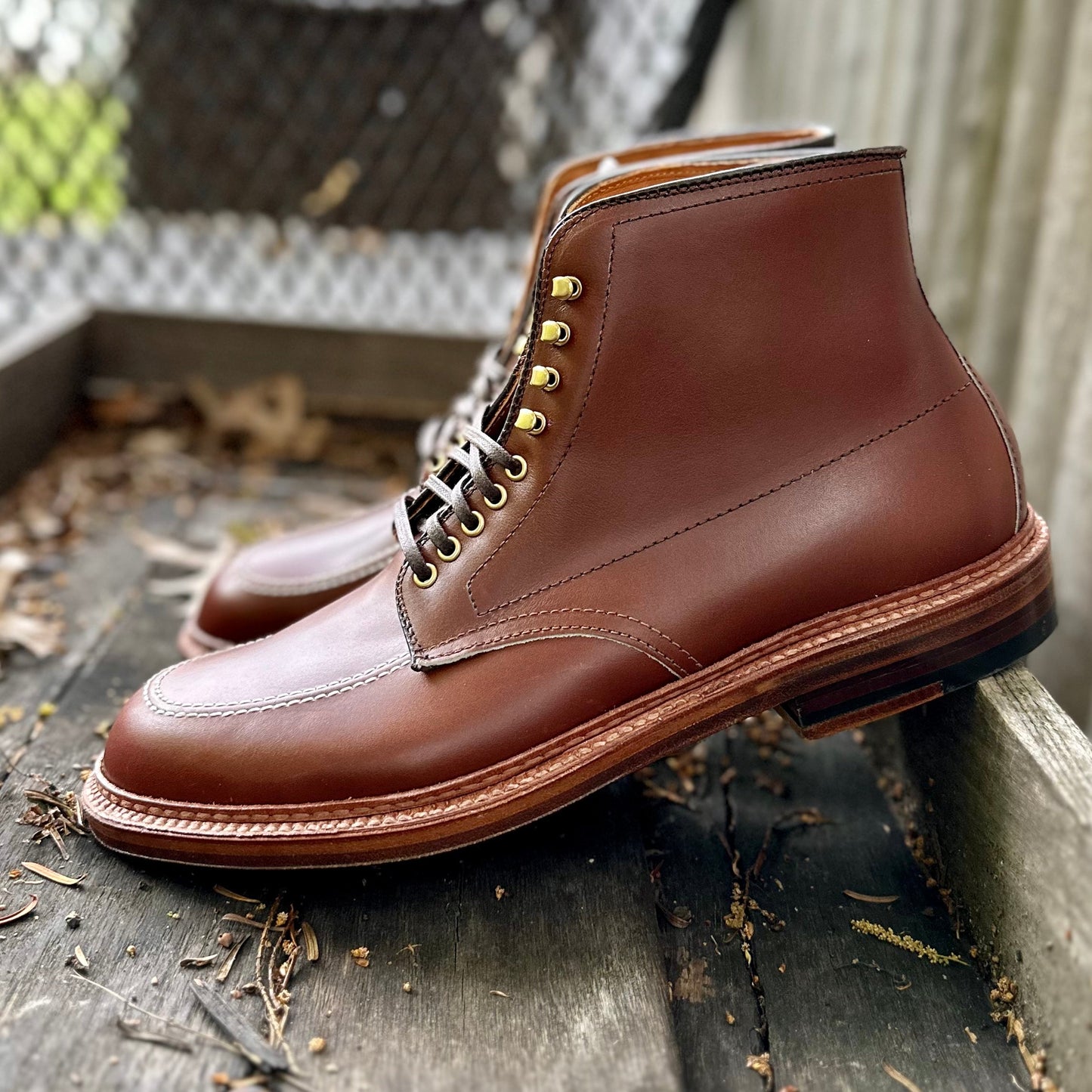 A pair of Alden Madisons D2940H - Madison Indy Boots in polished Madison Brown calf leather, featuring brass eyelets and a tan leather welt, sits on a weathered wooden surface outdoors. The rich calfskin complements the rugged scene with a chain-link fence and blurred greenery in the background.