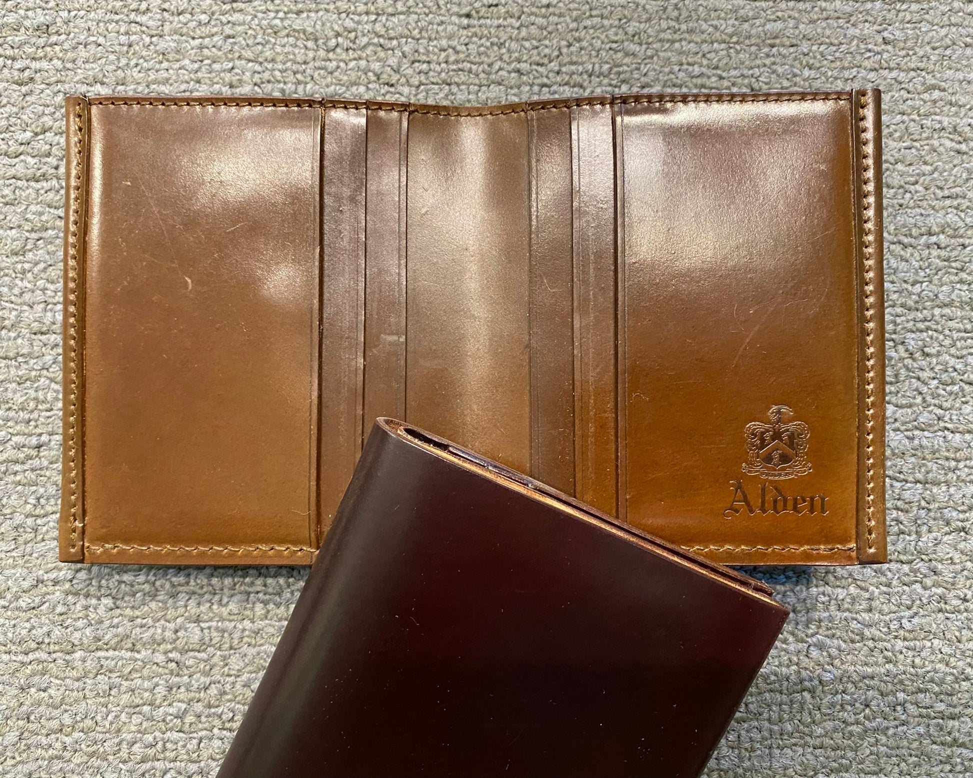 The LG 7007 - Alden Cognac Shell Cordovan Wallet by Alden Madison, embossed with the logo inside, rests on a textured surface. Crafted from Shell Cordovan leather, its artistry is remarkable. A dark brown wallet with sleek card slots peeks into the foreground.