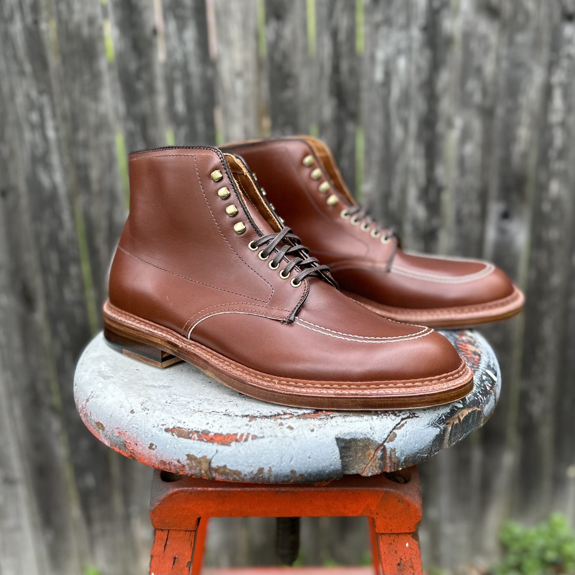 A pair of D2940H - Madison Indy Boots in Madison Brown Calf by Alden Madison, featuring white stitching and metal eyelets, rests on a paint-splattered stool in front of a gray wooden fence.