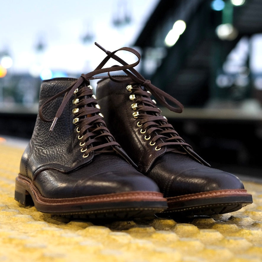 The Alden Madison D1808HC - Stitchup 2/2 Boot in Arabica Lux7 features dark brown calfskin leather, gold eyelets, dark laces, and a sturdy Commando Sole, displayed on a textured yellow surface outdoors with blurred lights in the background.