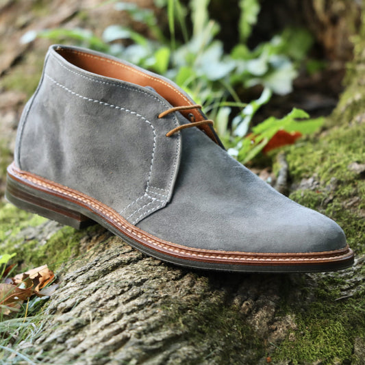 The Alden Madison 1592L Chukka Boot with L3 Sole in Lead Suede, featuring brown laces and a Reverse Antique Welt, sits atop a mossy tree trunk outdoors amid lush green foliage.