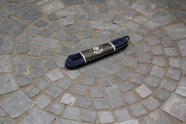 A coiled 27-inch Alden Madison Guarded Goods shoe lace rests at the center of a circular stone-tiled ground, wrapped in a paper label.
