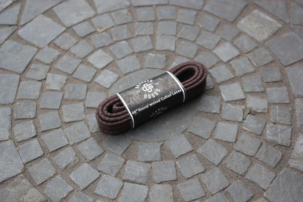 A pair of Alden Madison Laces - Round Boot Laces - 48 Inch, brown with a black and white label, is coiled on a circular stone tile pattern.