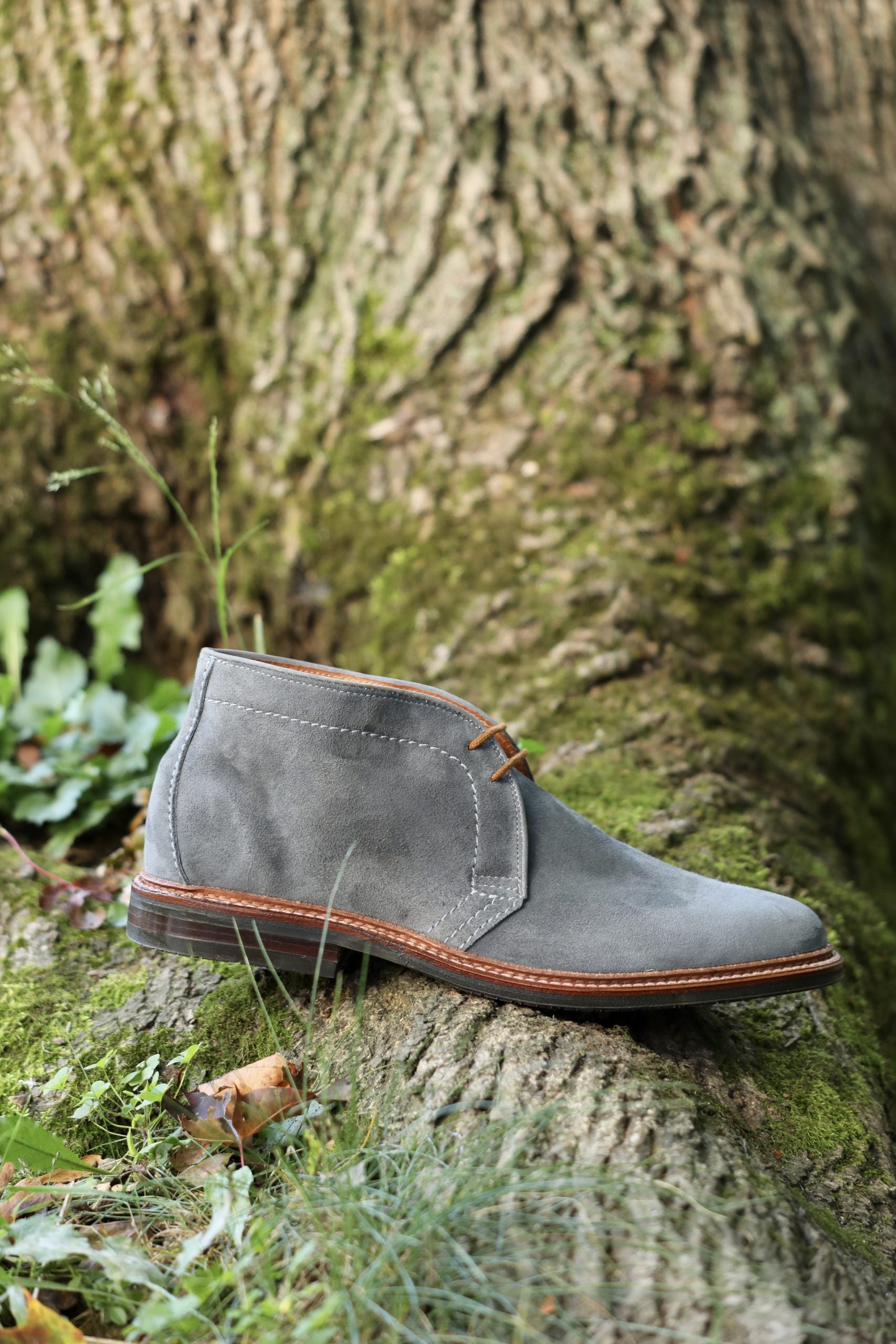 The Alden Madison 1592L Chukka Boot in lead suede with an L3 sole sits on mossy tree bark, surrounded by grass and leaves outdoors.